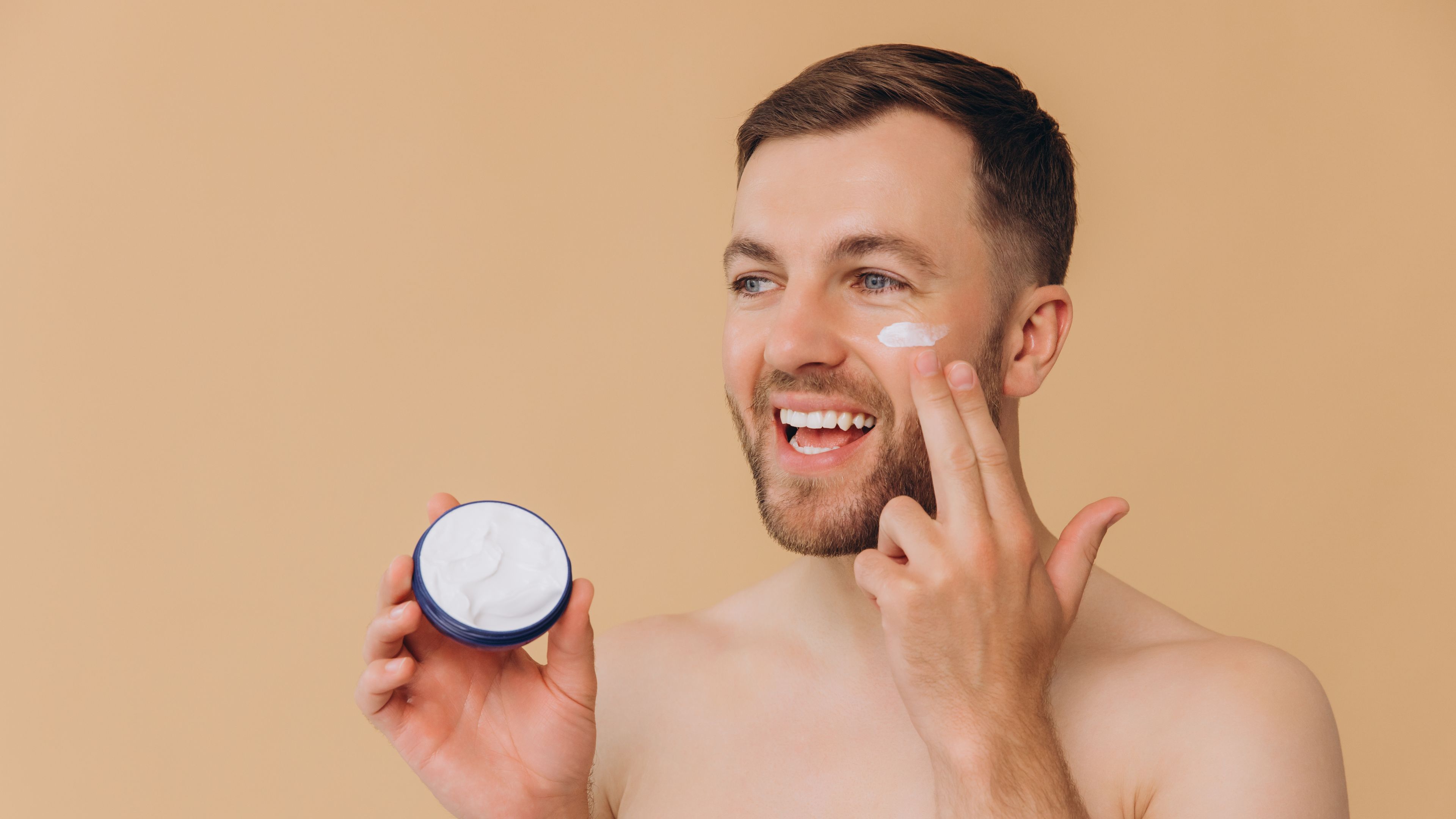 Attractive happy smiling bearded man with healthy skin, applies cream moisturizer for anti wrinkle.