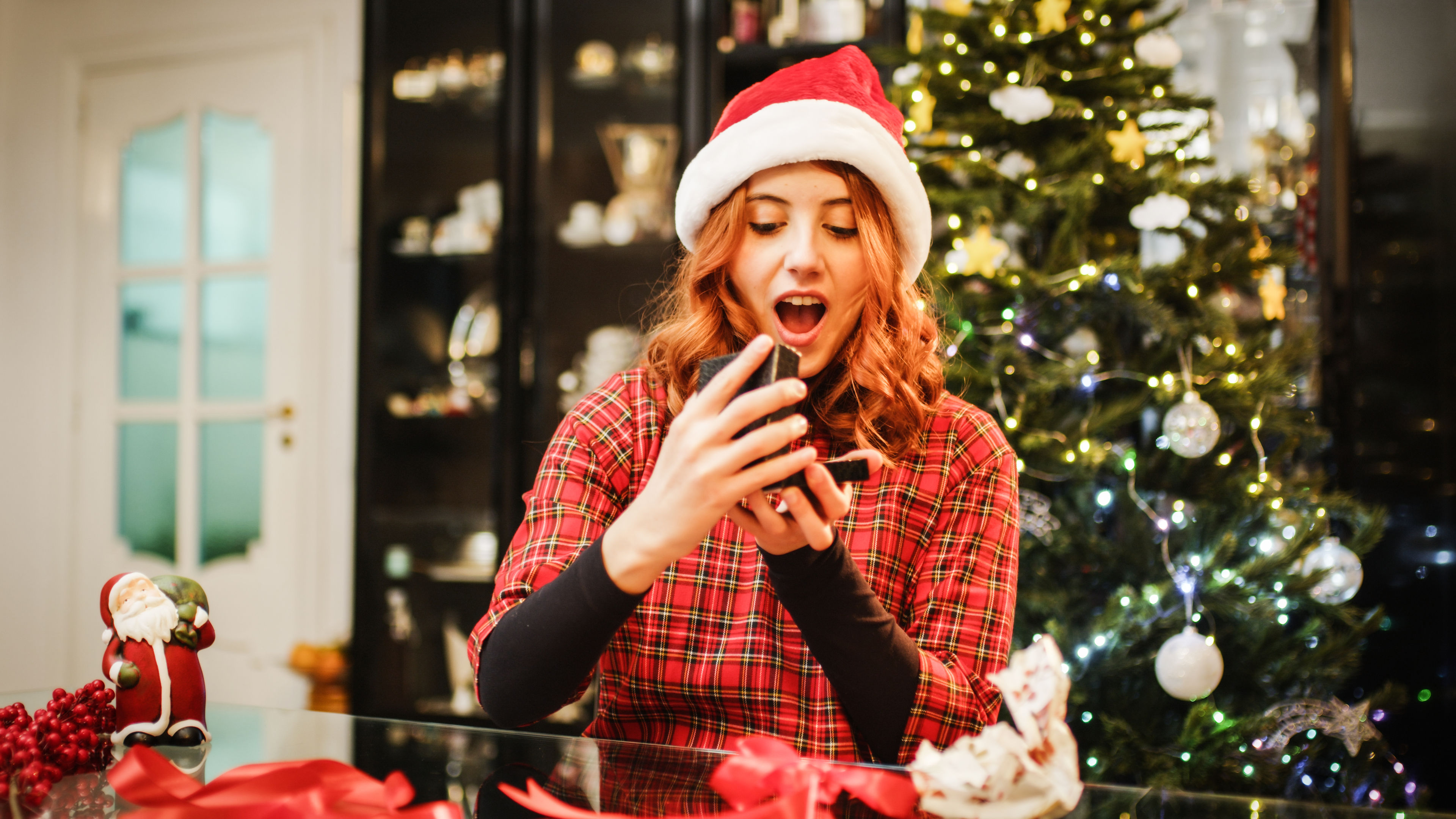 A young girl in a festive Santa hat excitedly opens a beautifully wrapped gift by a glowing Christmas tree 