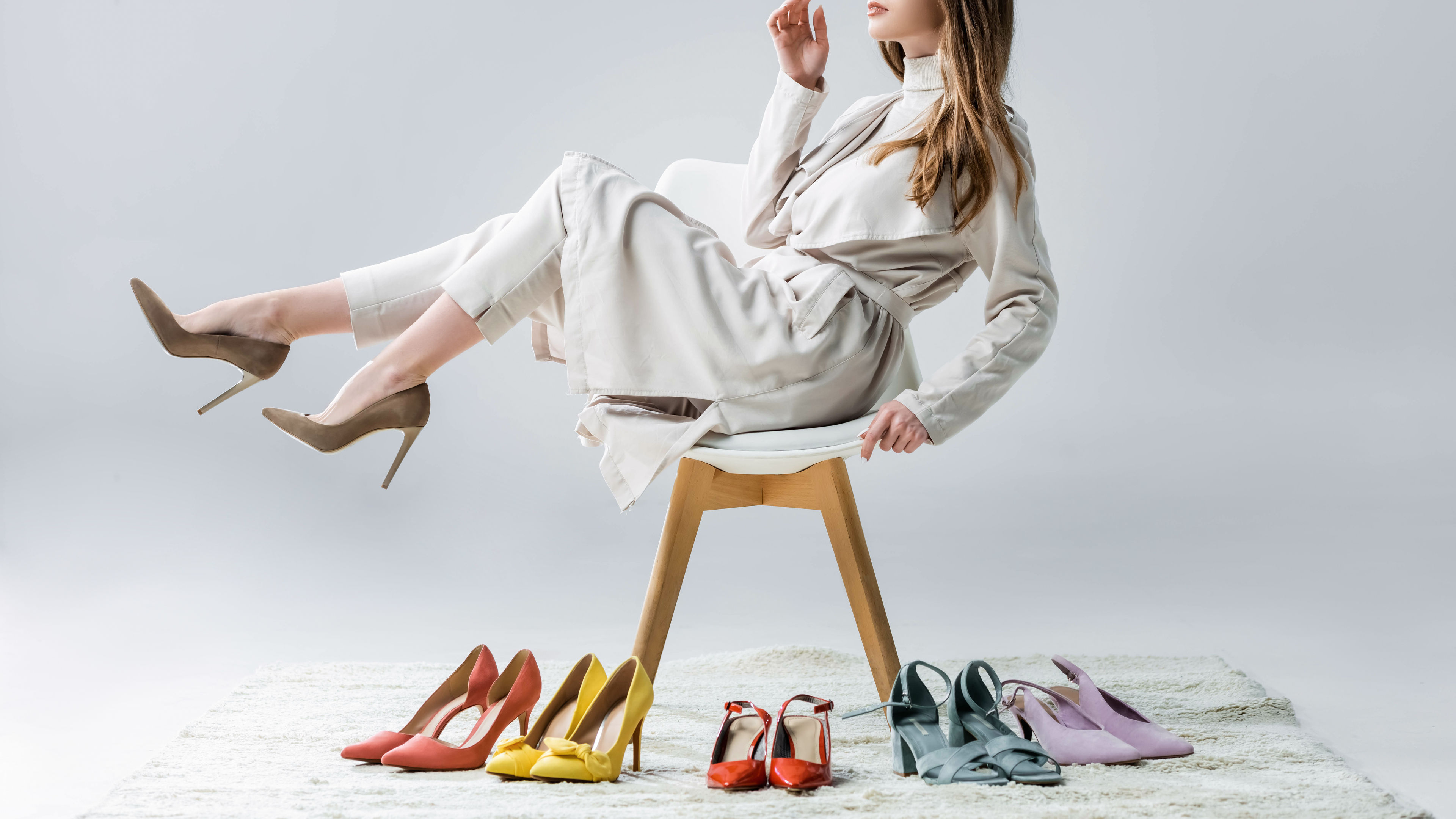Thoughtful girl sitting on a chair with raised legs near a collection of shoes and looking away on the grey.