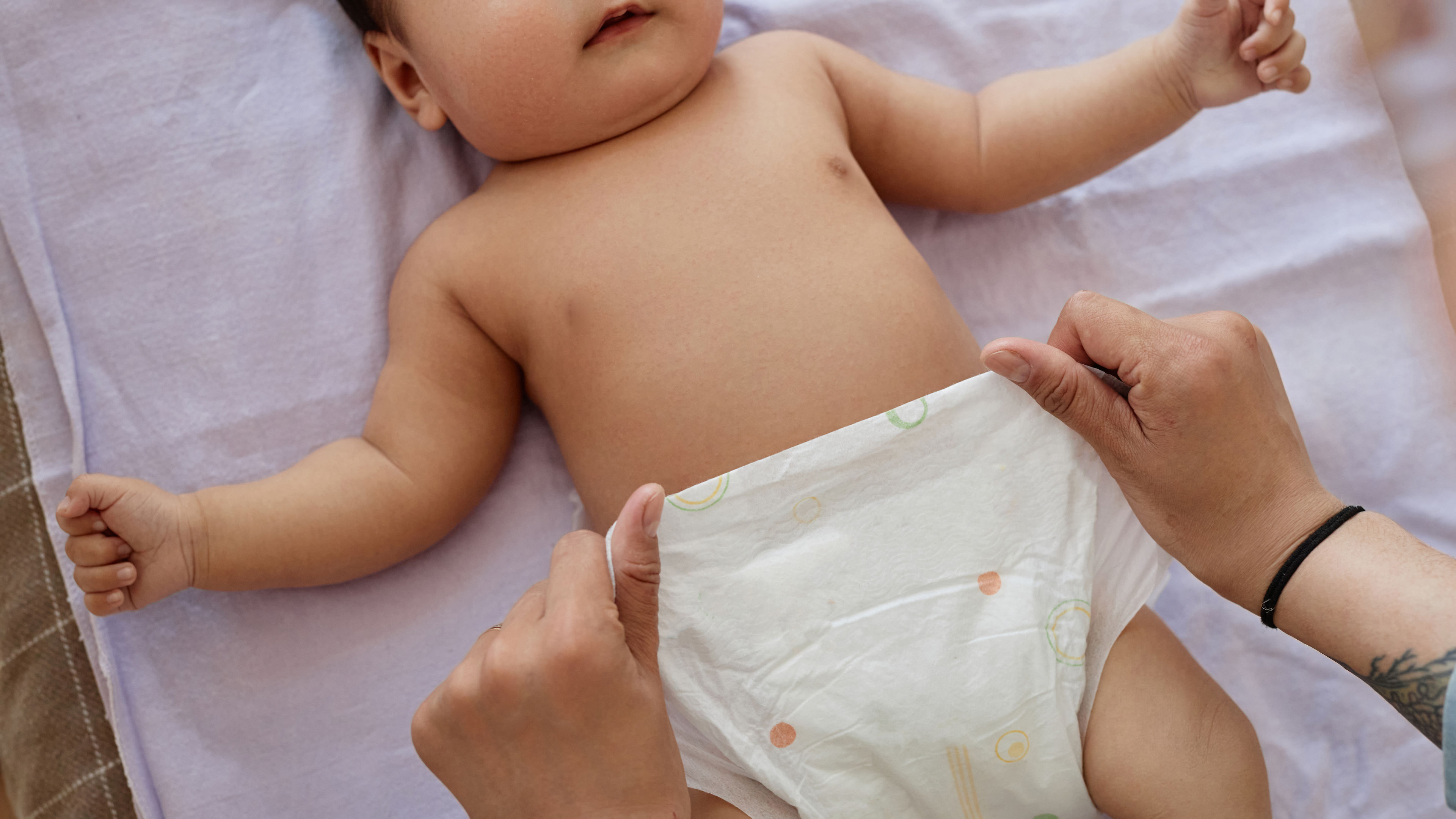 A baby is lying on the bed, smiling, while its mom is putting on a diaper.