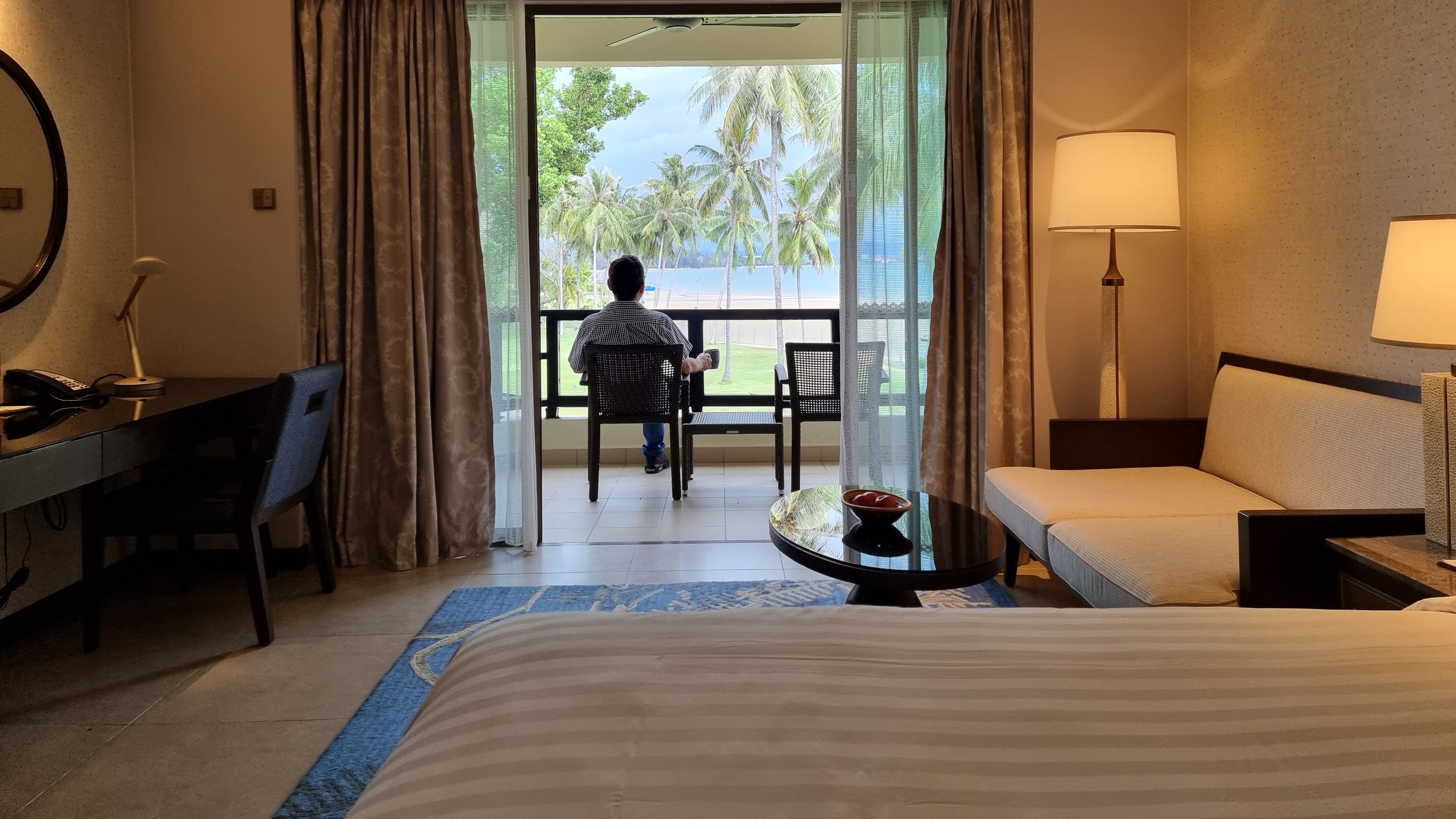 A man is comfortably seated on the balcony of a hotel room, enjoying the view and the warm sunlight.