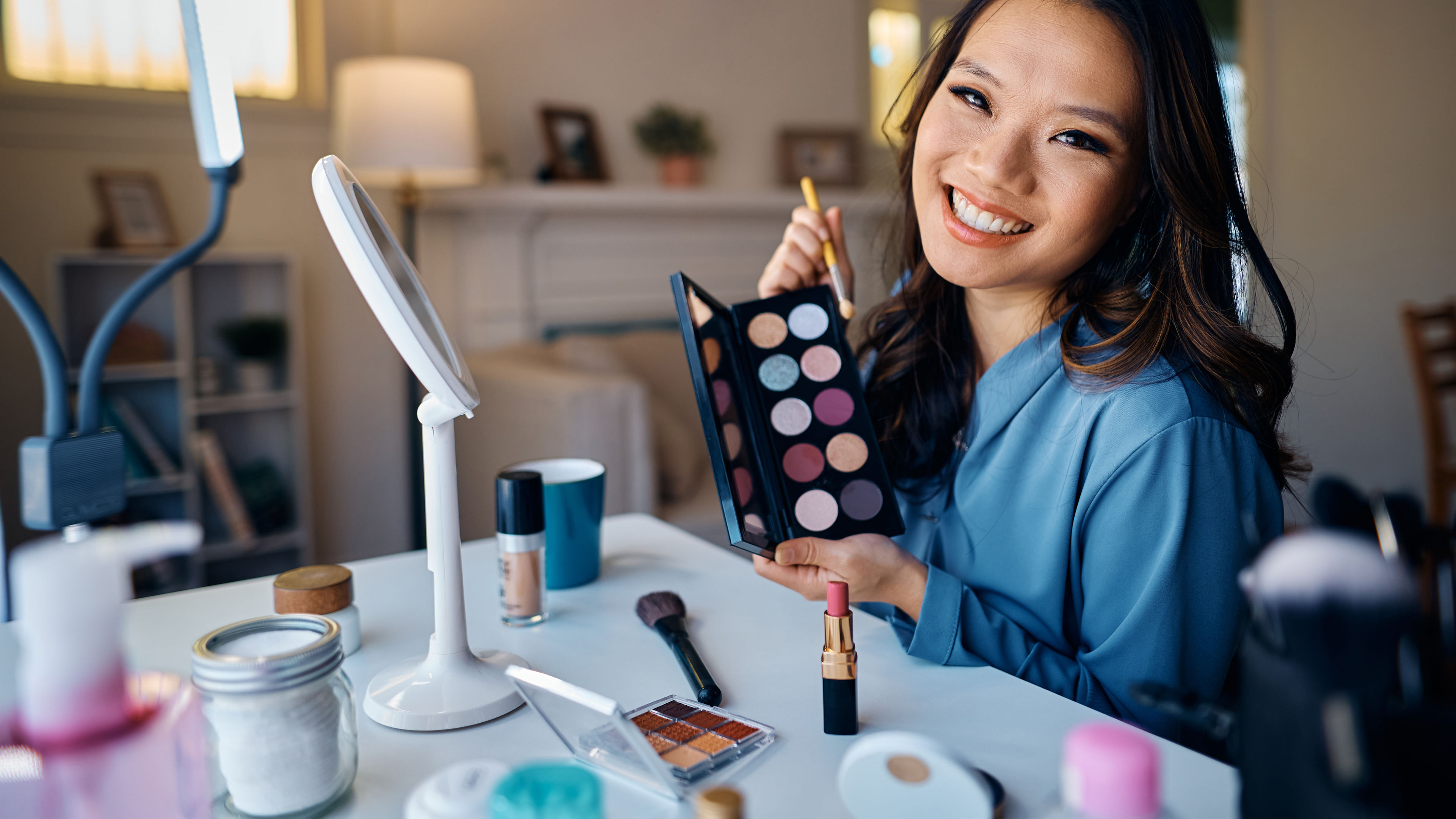 A woman smiling while showcasing an eyeshadow palette, seated at a makeup station with various cosmetics.