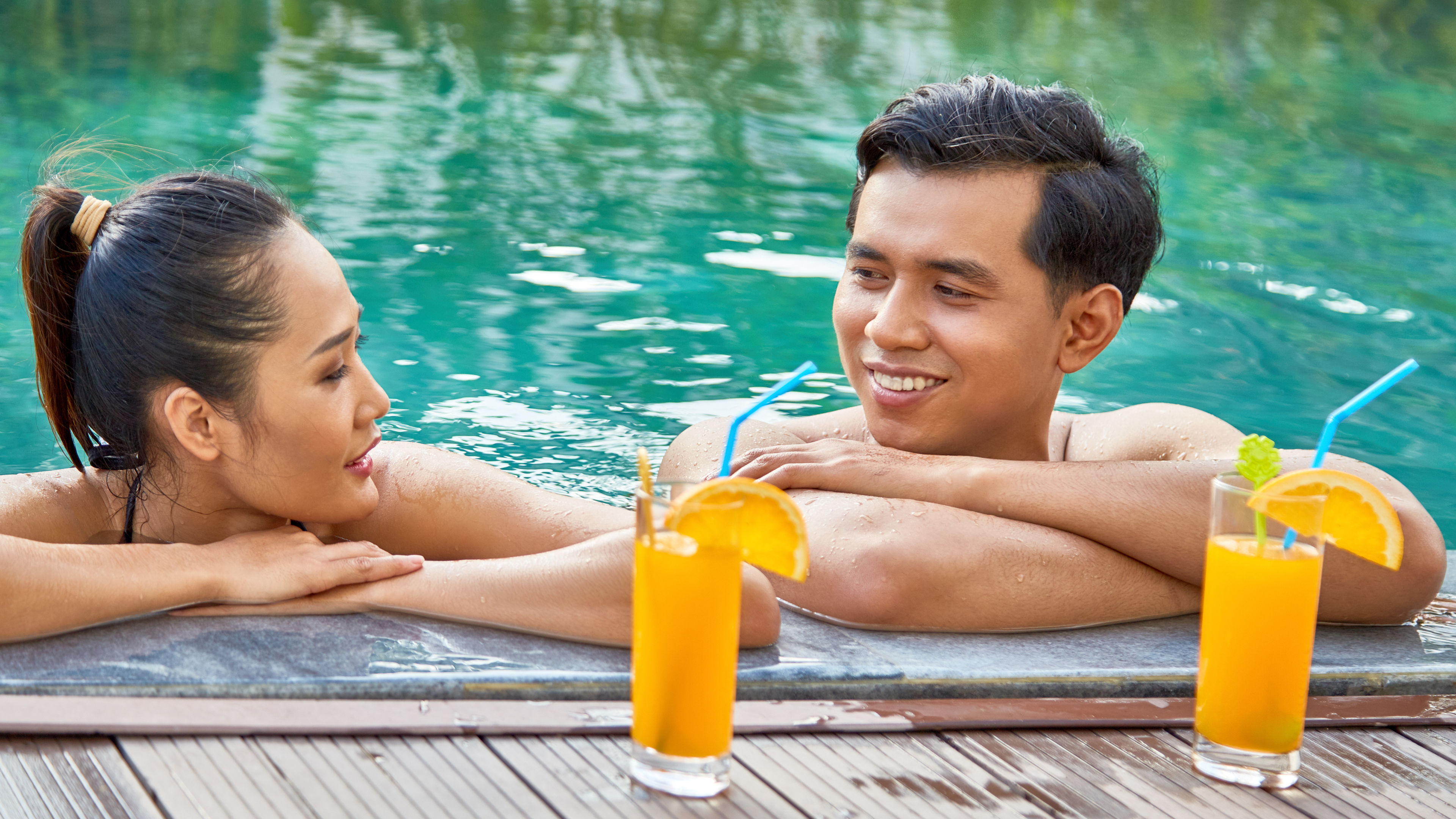 A happy couple relaxing in a swimming pool with glasses of orange juice placed nearby.