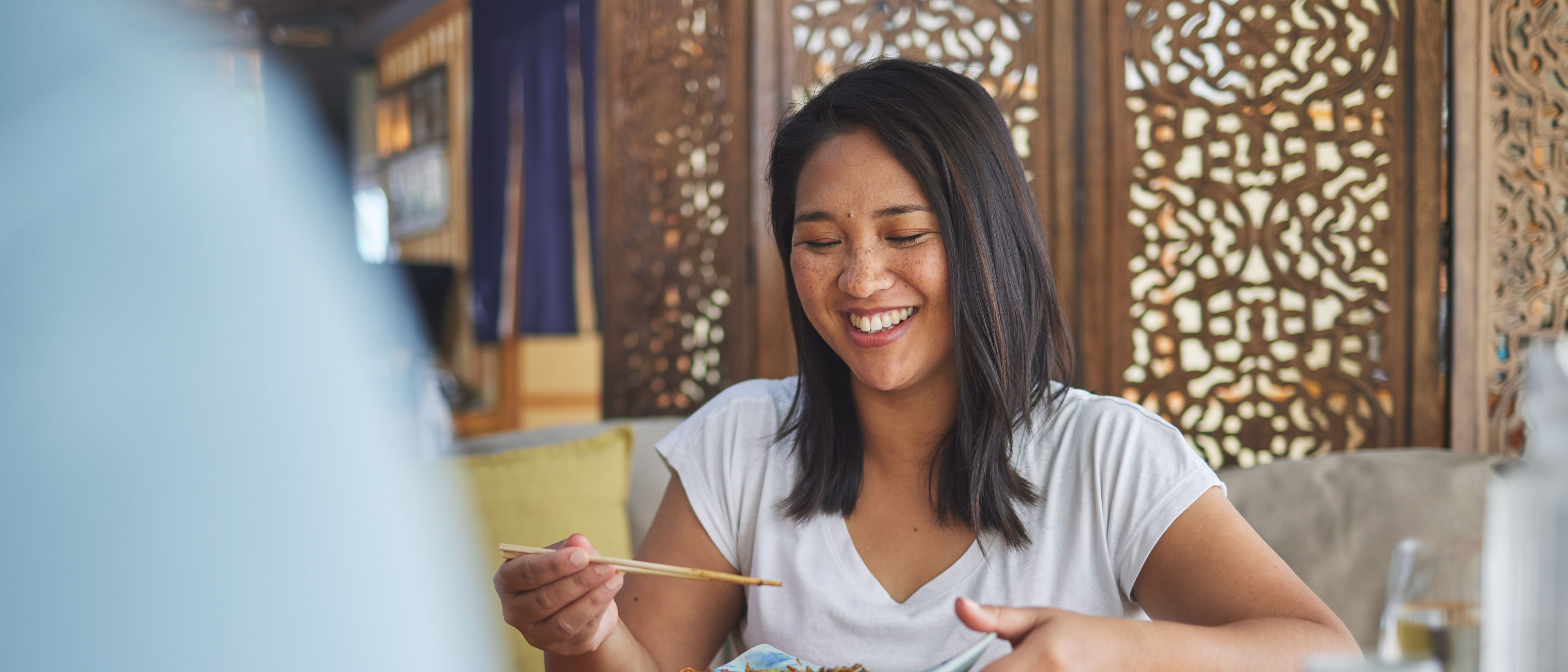 
A happy woman is eating food at a restaurant, smiling as she enjoys her meal.