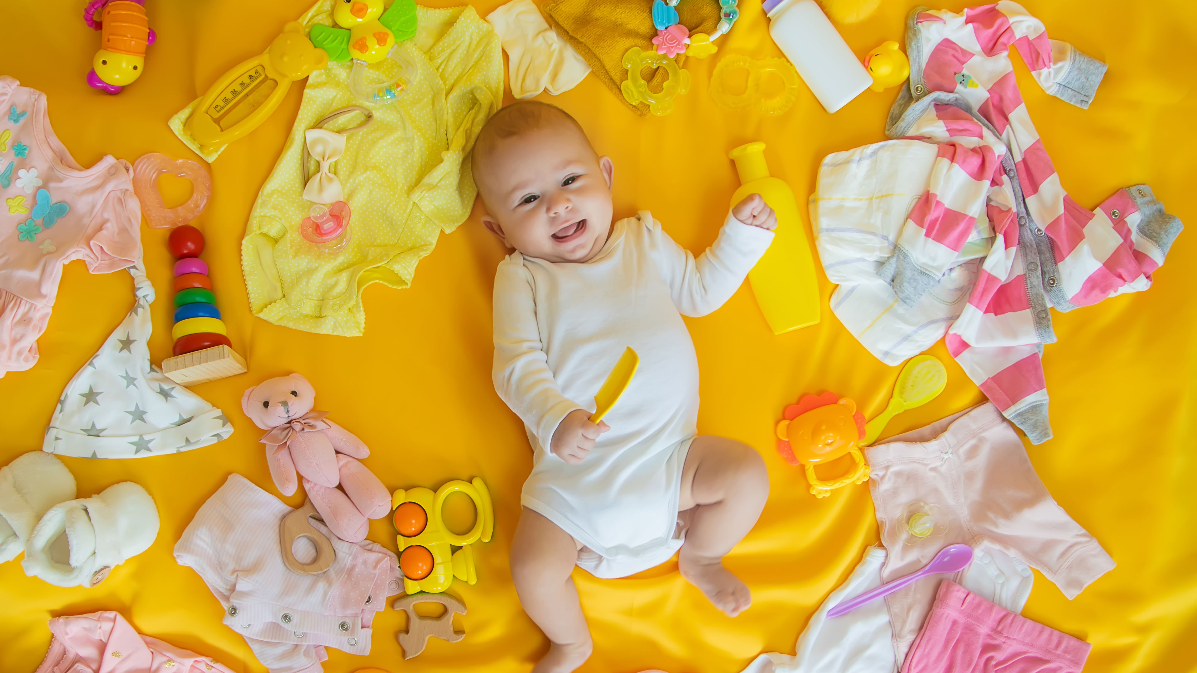 A happy baby lying on a yellow surface, surrounded by colorful toys and dresses.