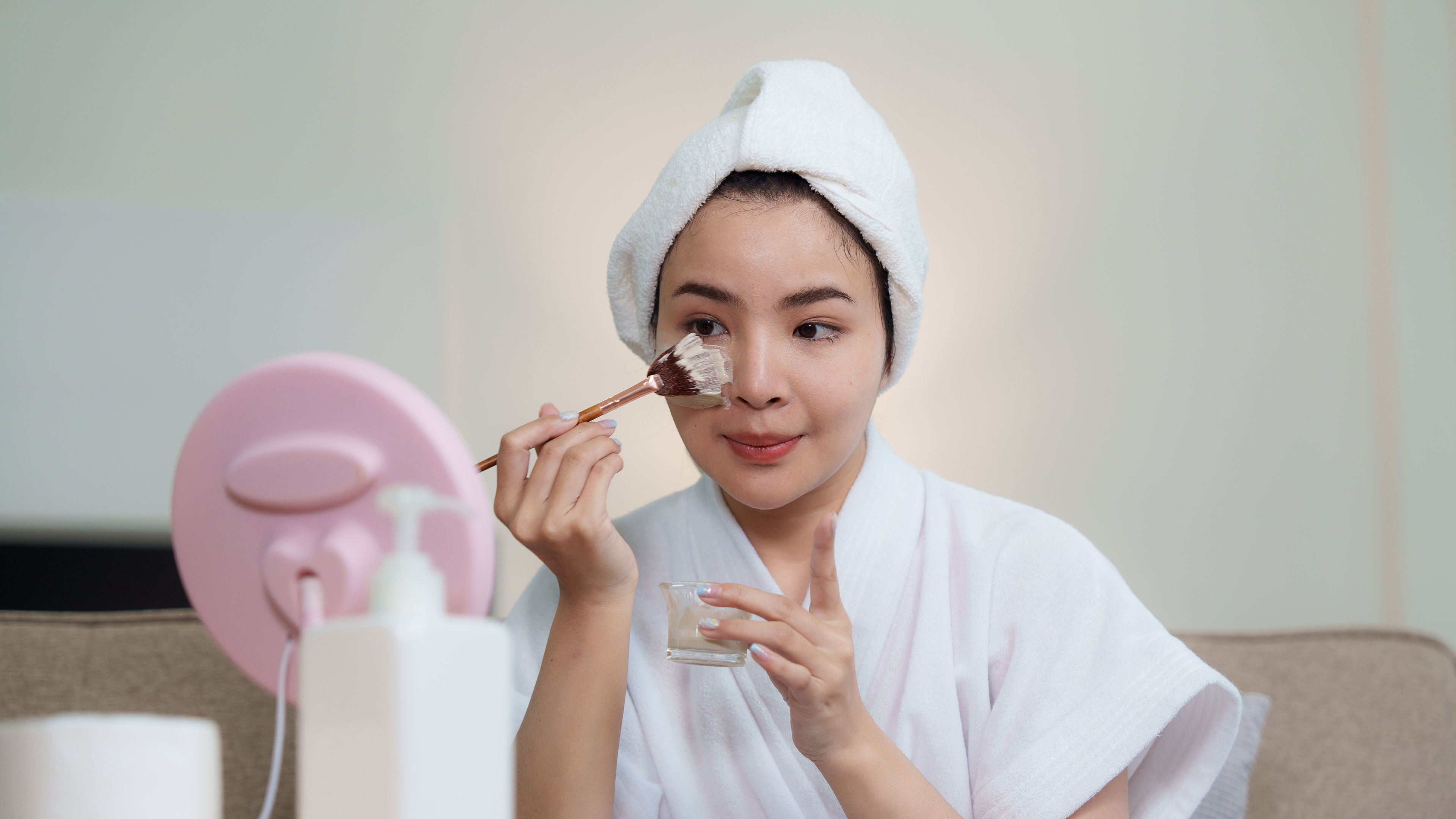 穿着浴袍嘅女人喺鏡前用刷子敷面膜。