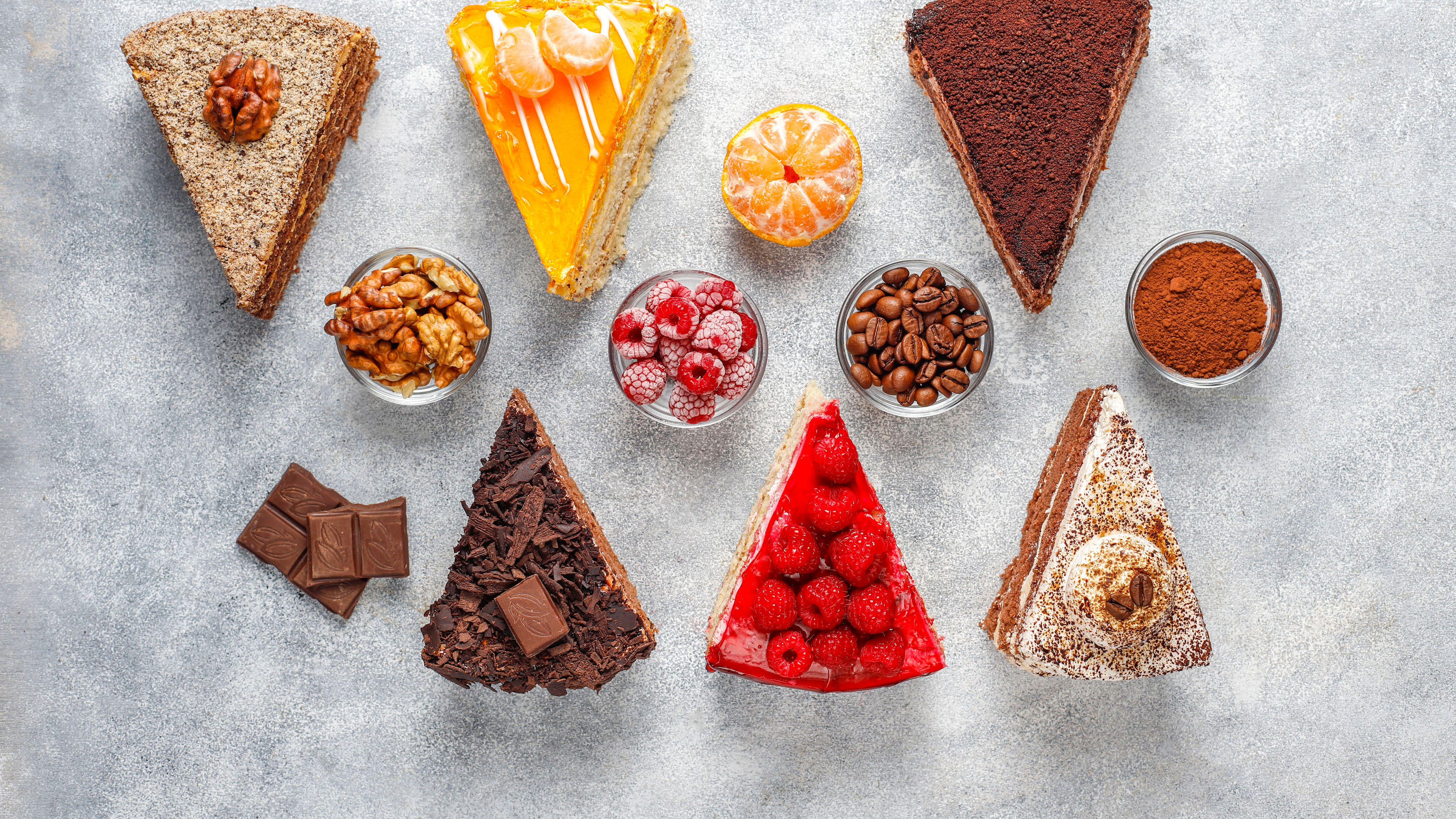 Various colorful cake slices topped with fruits, chocolate, and nuts, beautifully displayed with matching ingredients. 