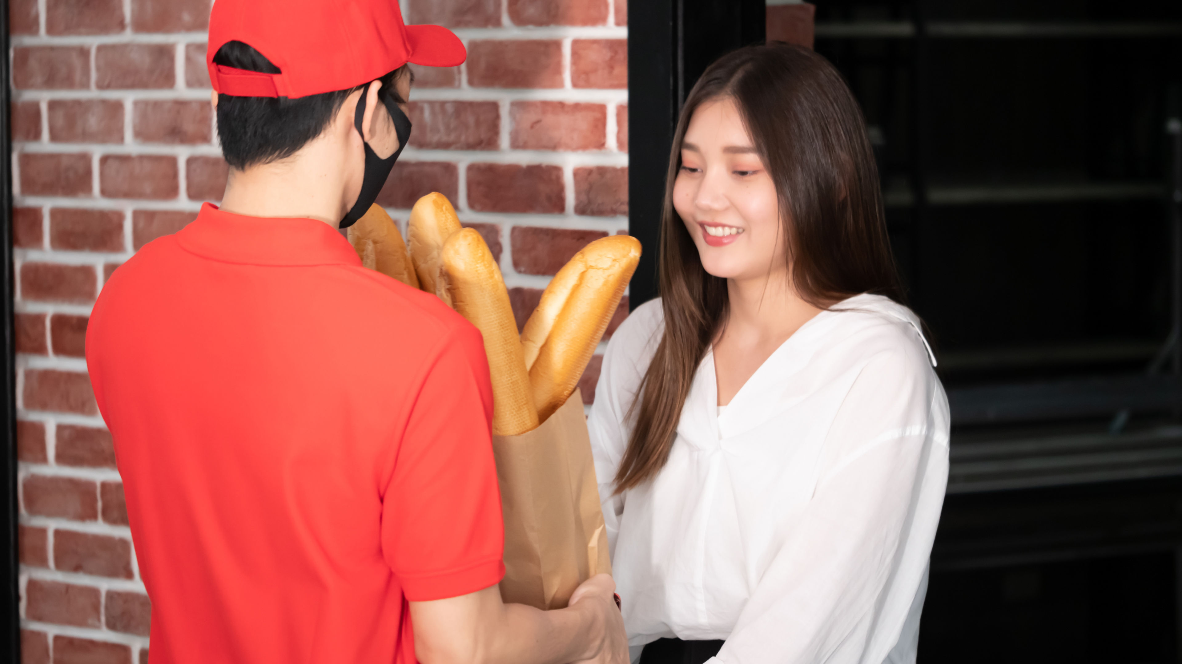 A woman warmly receives a paper bag filled with baguettes from a delivery person 