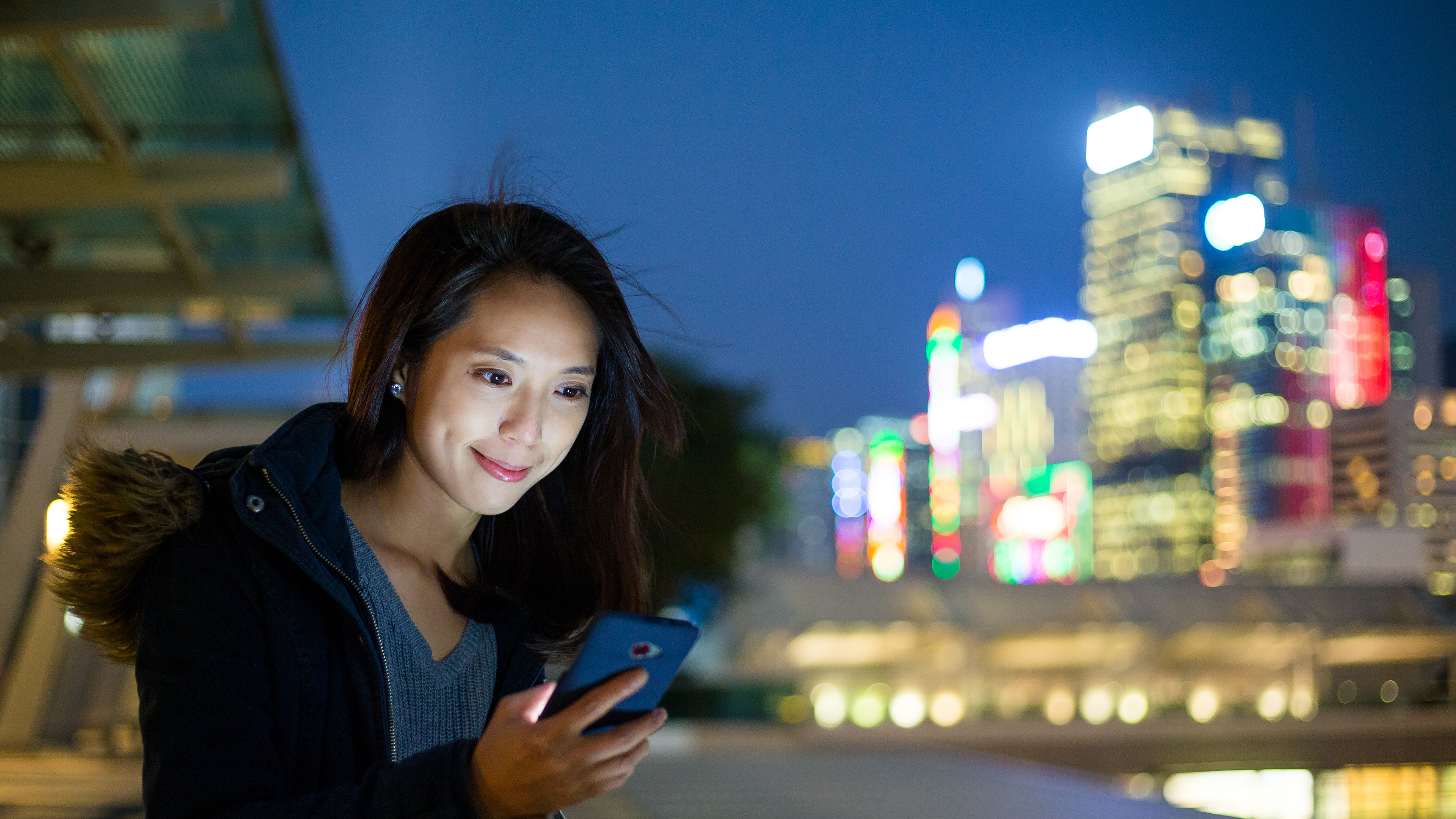 A young woman stands in the vibrant streets of Hong Kong, engrossed in her cellphone. 