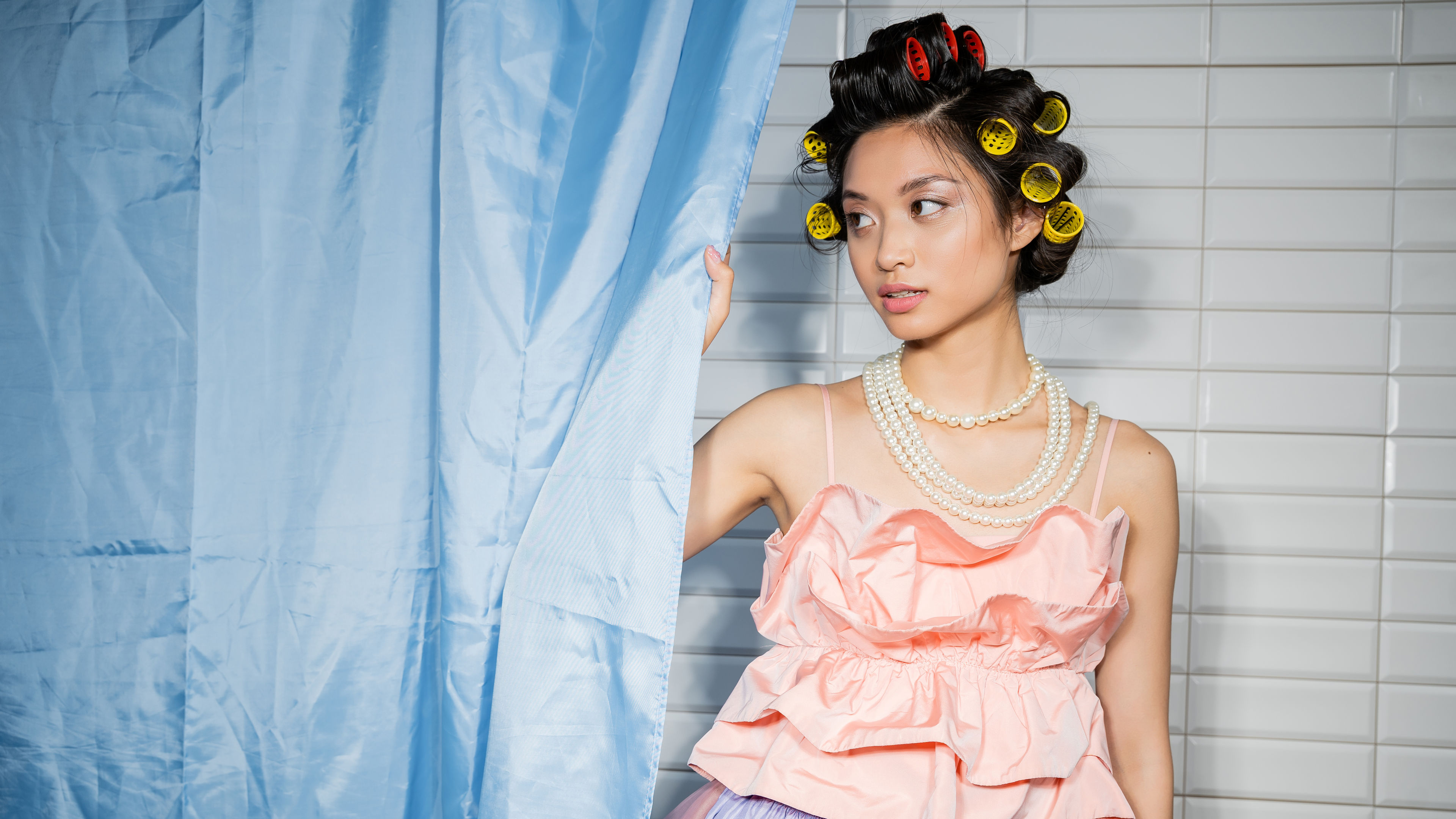 alluring and asian young woman with hair curlers standing in pink ruffled top with pearl necklace 