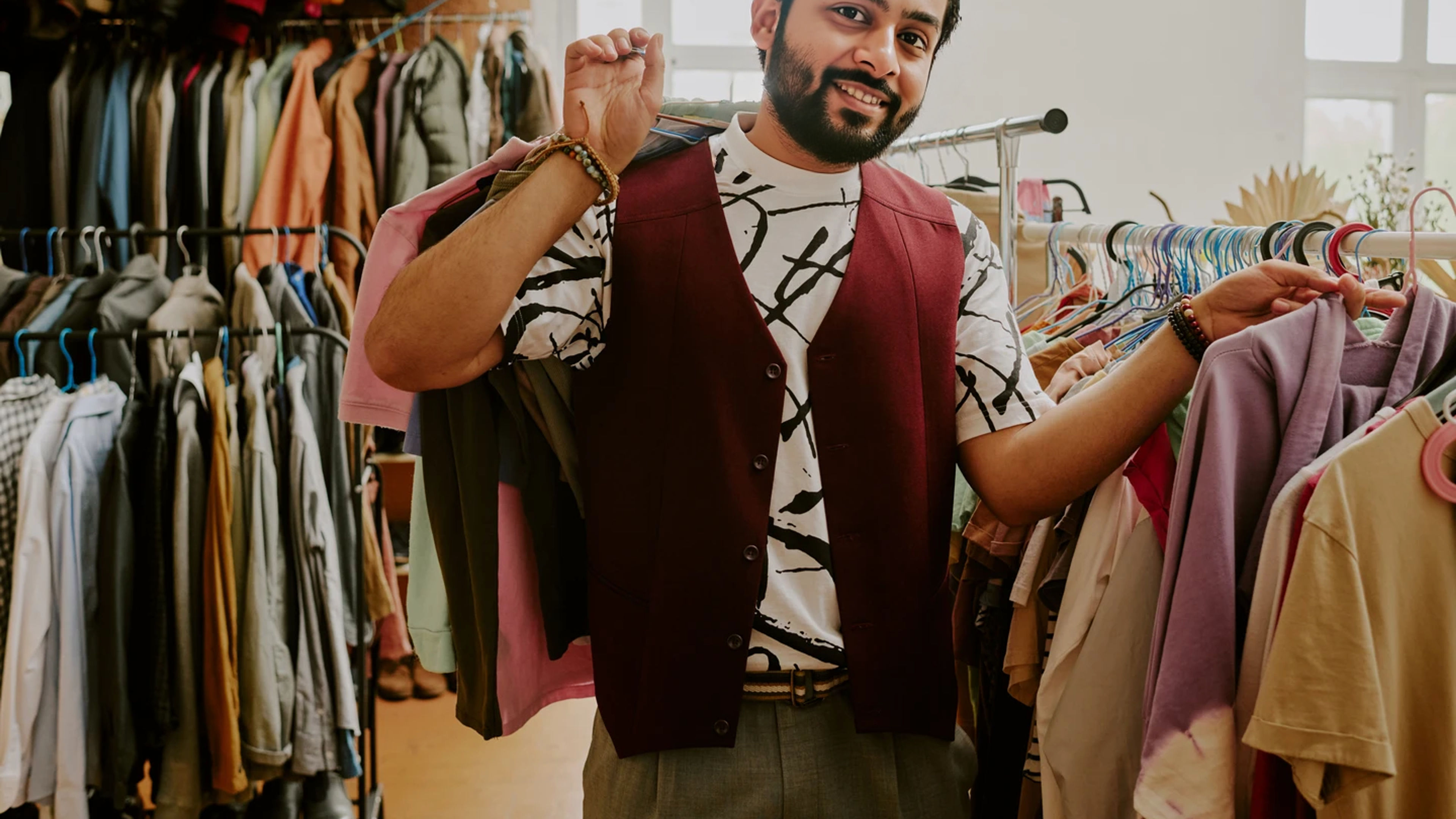 Smiling man shopping in a clothing store, surrounded by various garments on hangers, holding a jacket over his shoulder and enjoying the experience.
