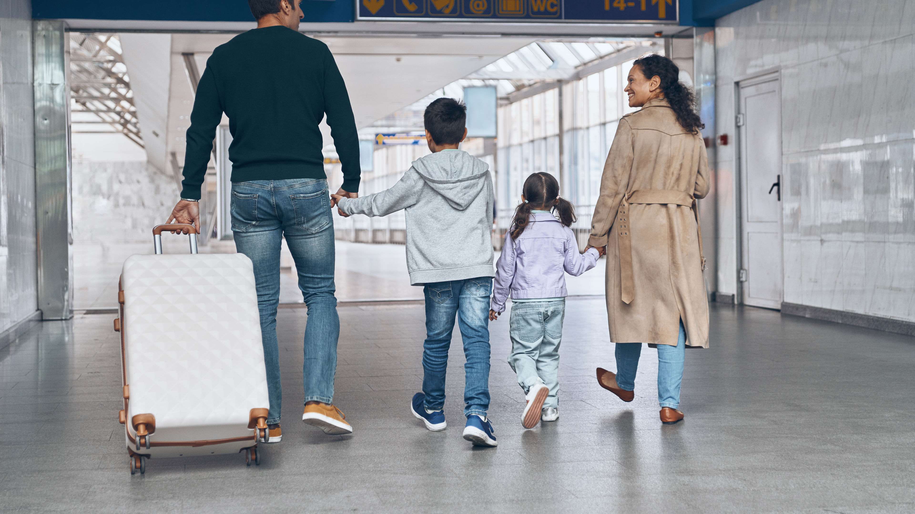 Rearview of a happy Indian family with a white trolley bag in an airport.