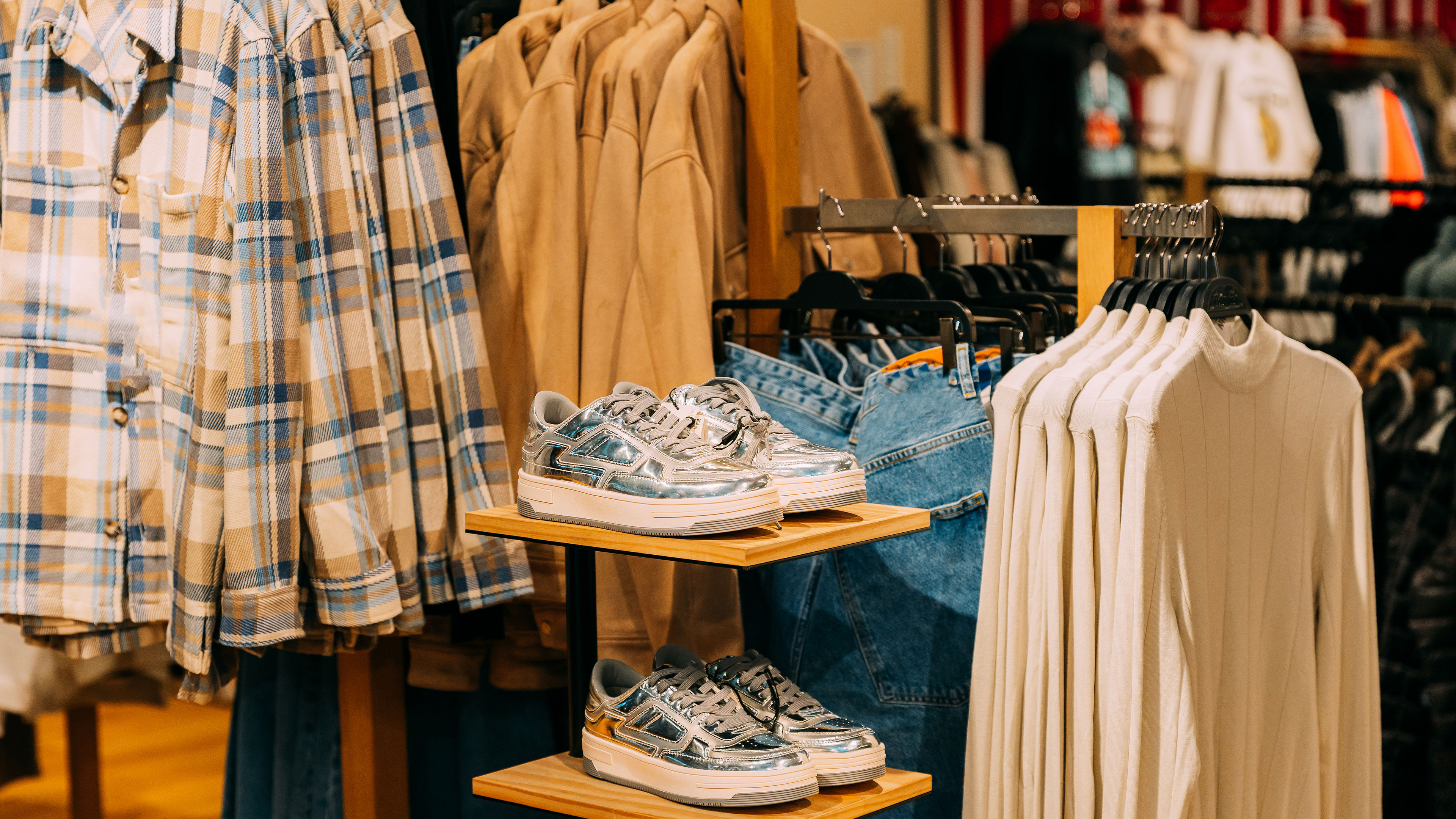 Silver unisex sneakers displayed on a shelf with clothes hanging in the background in a mall store.
