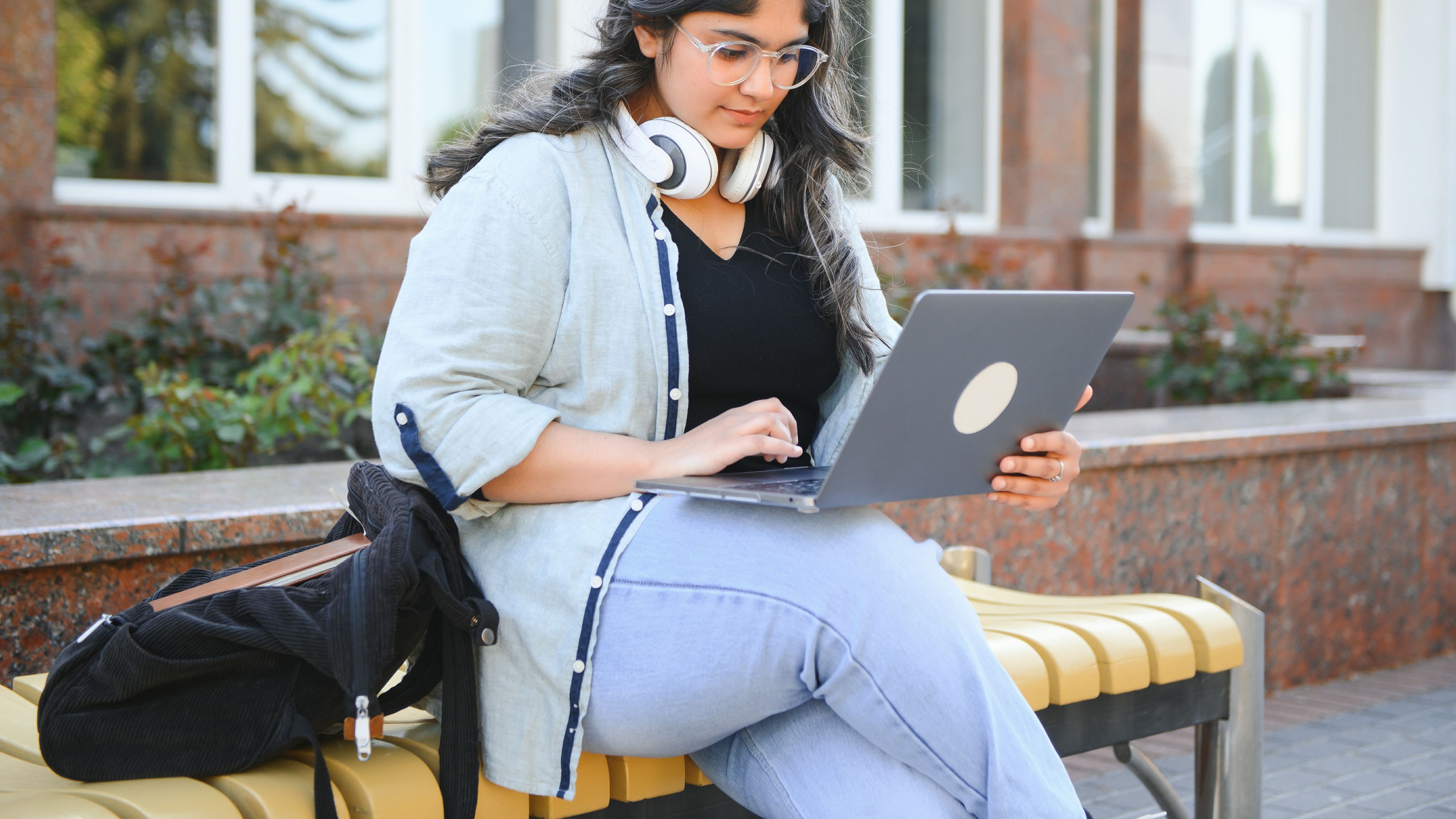 A young student wearing headphones and glasses sits on a bench outdoors, working on her laptop 