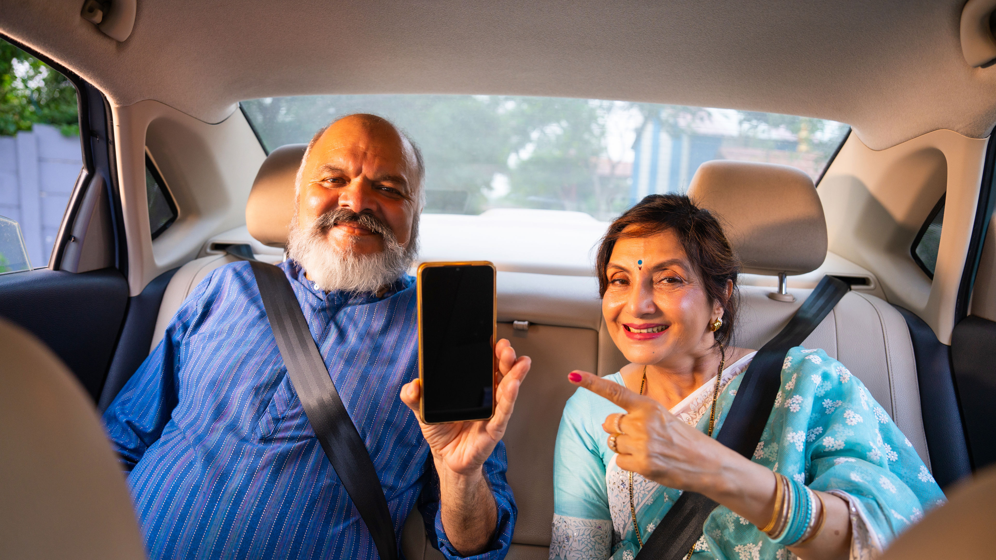 Senior Indian couple, seated in a luxury car back seat, showcasing a smartphone with a blank screen 