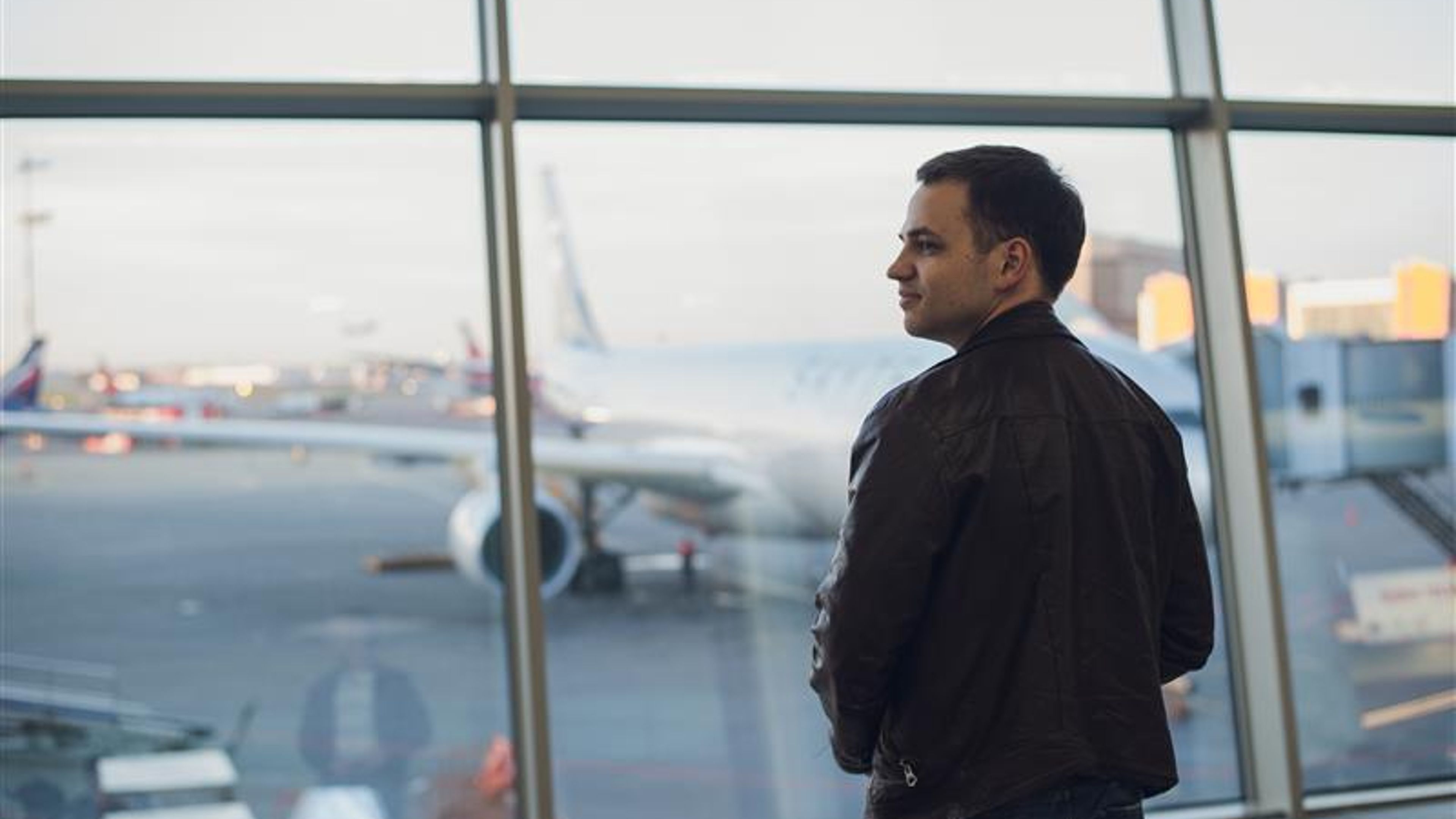 Young man is standing near window at the airport and watching plane before departure. 