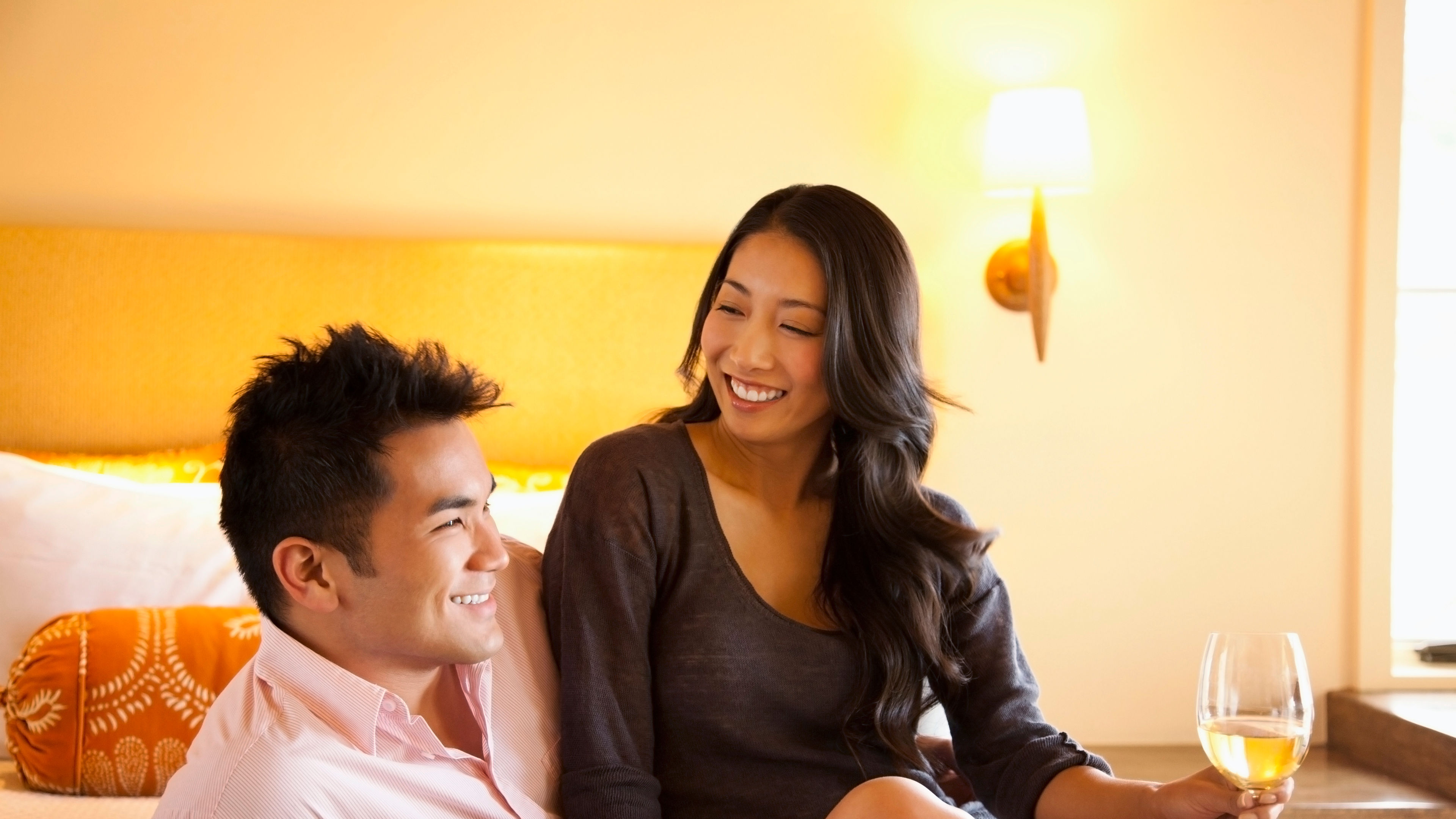 A couple sitting together on a bed smiling at each other warmly the woman holding a glass of wine.