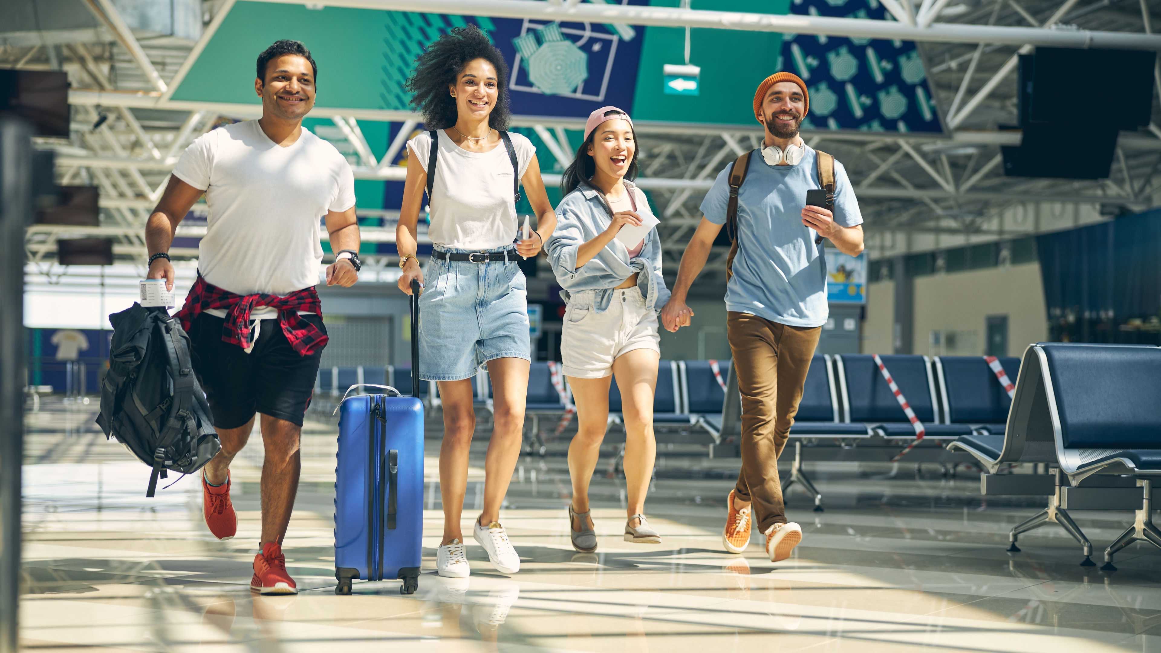 Four friends running through the airport with smiles on their faces, each carrying their own piece of luggage.