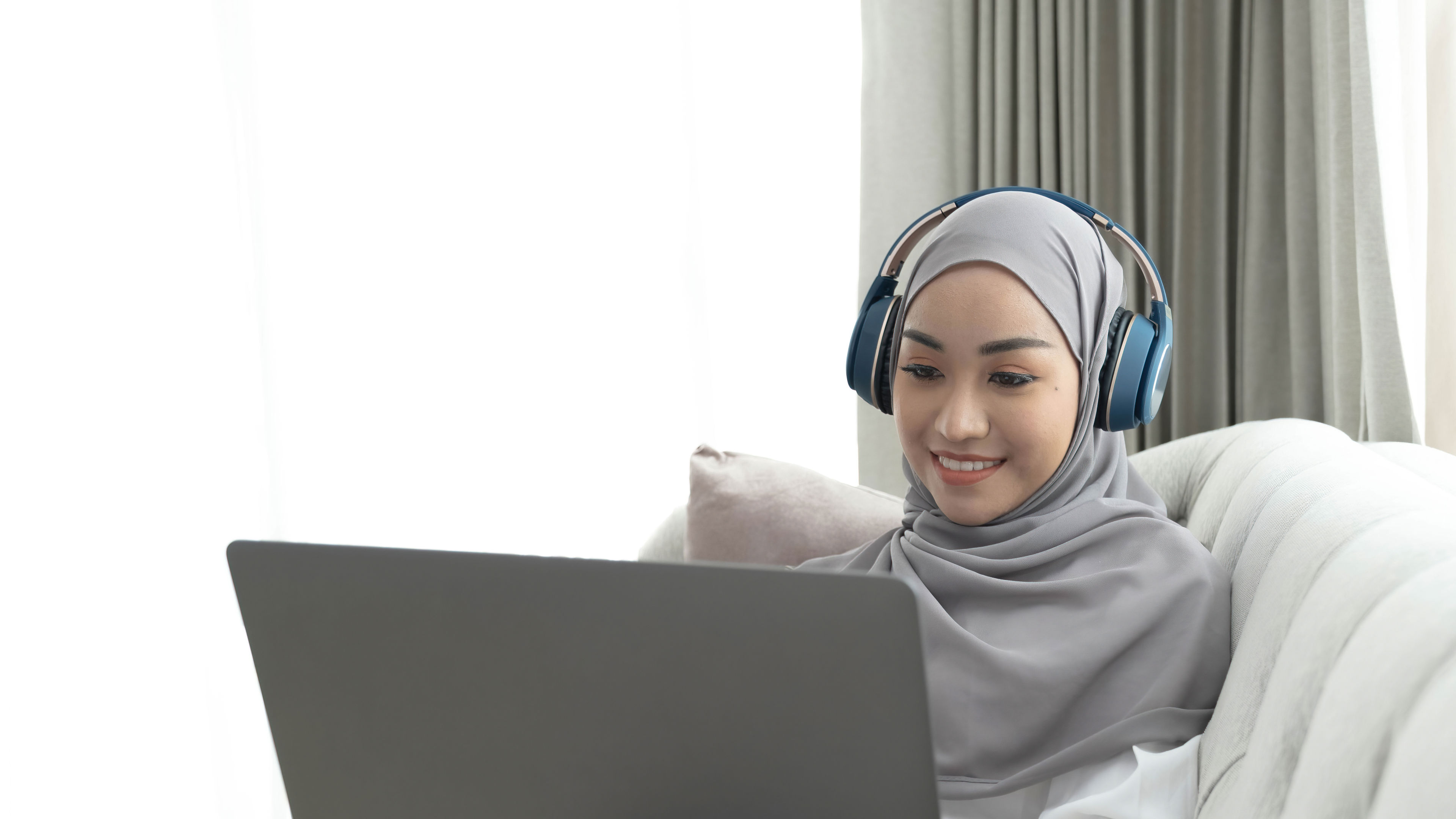 Wanita muda Muslim Asia yang menawan memakai tudung duduk di sofa yang selesa, menggunakan komputer riba 