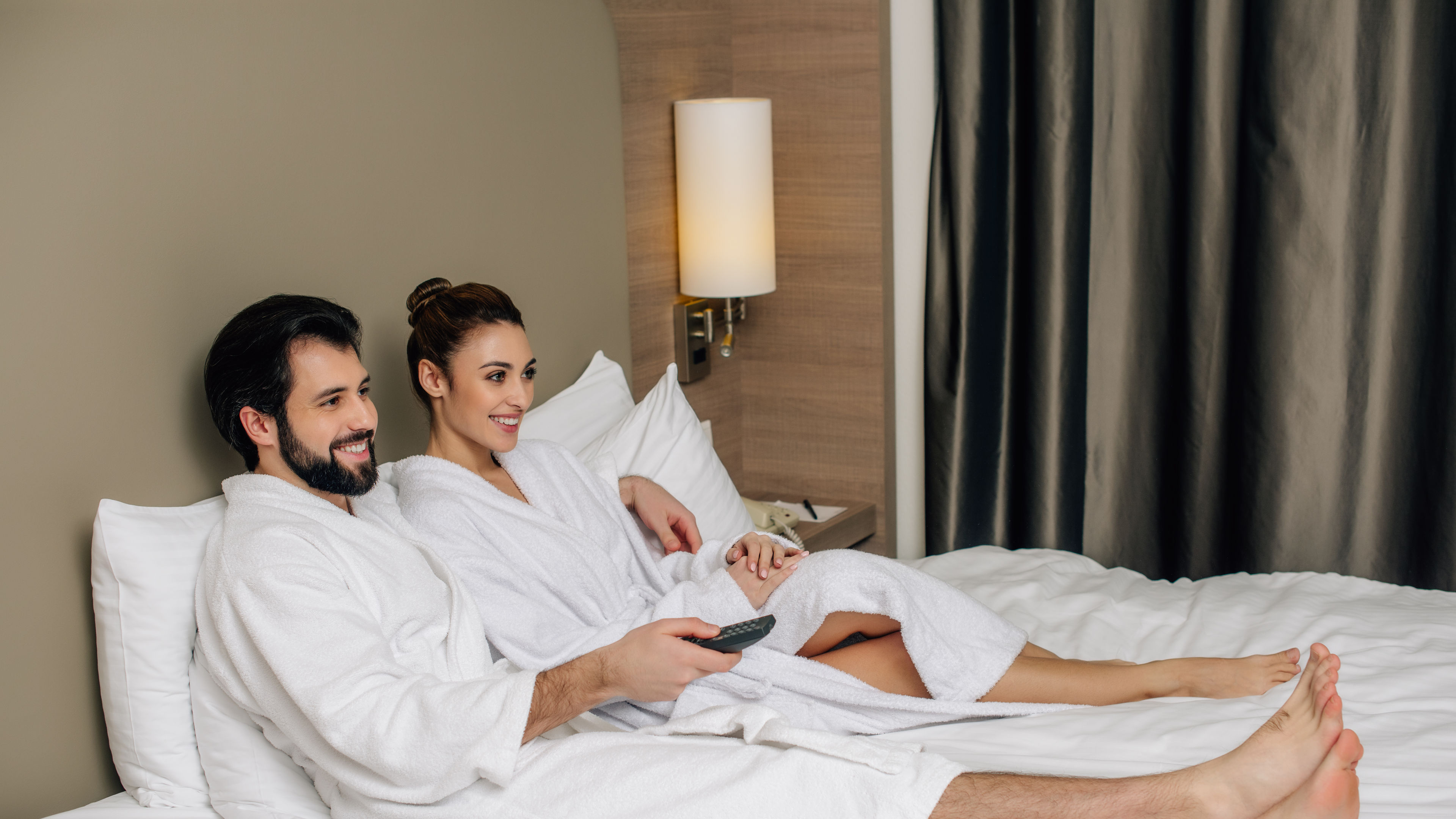 Beautiful couple in bathrobes with remote control relaxing in bed at hotel suite and watching tv.