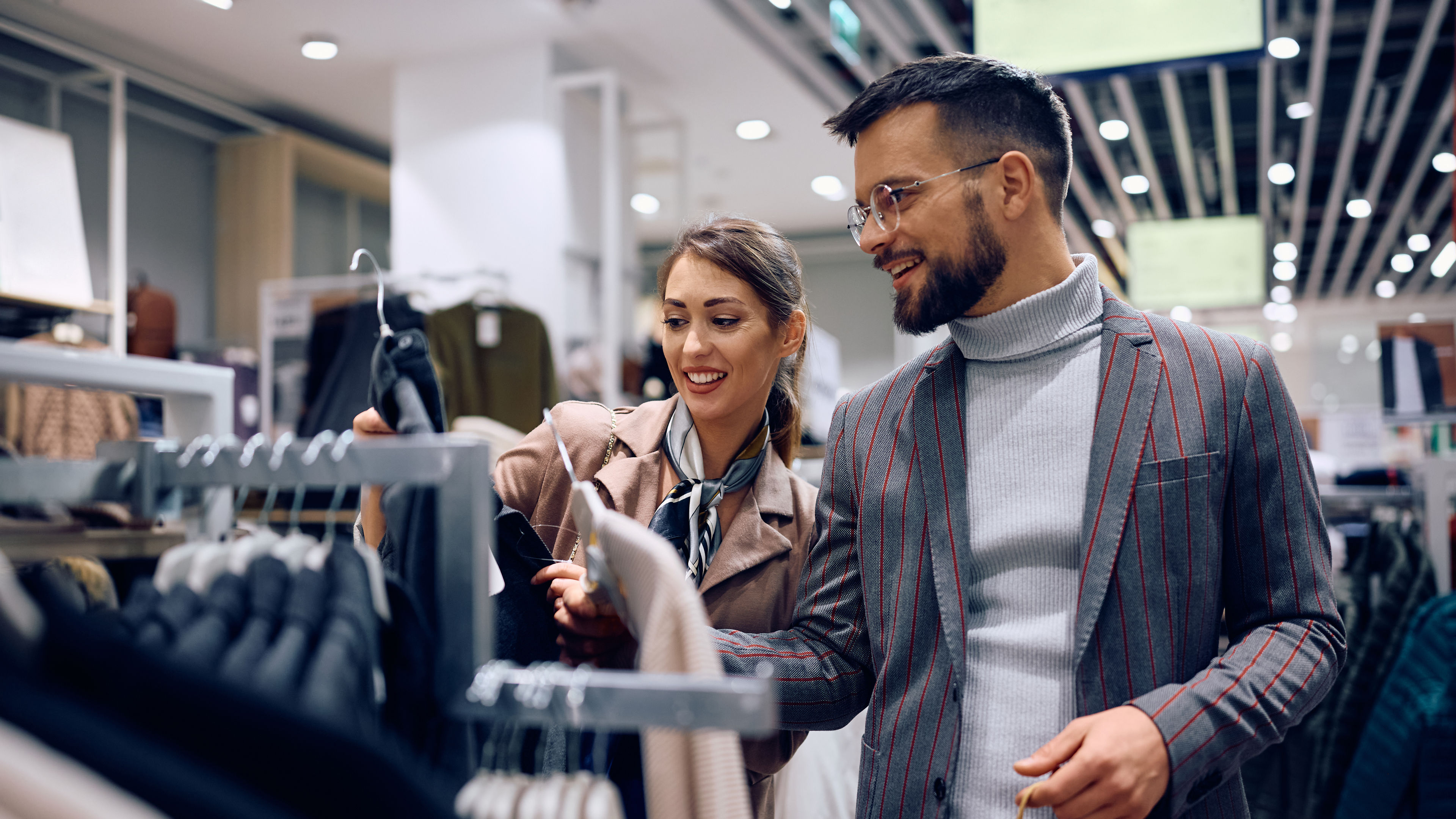 A man and a woman shopping for clothes in a store, browsing sweaters on racks with smiles on their faces.
