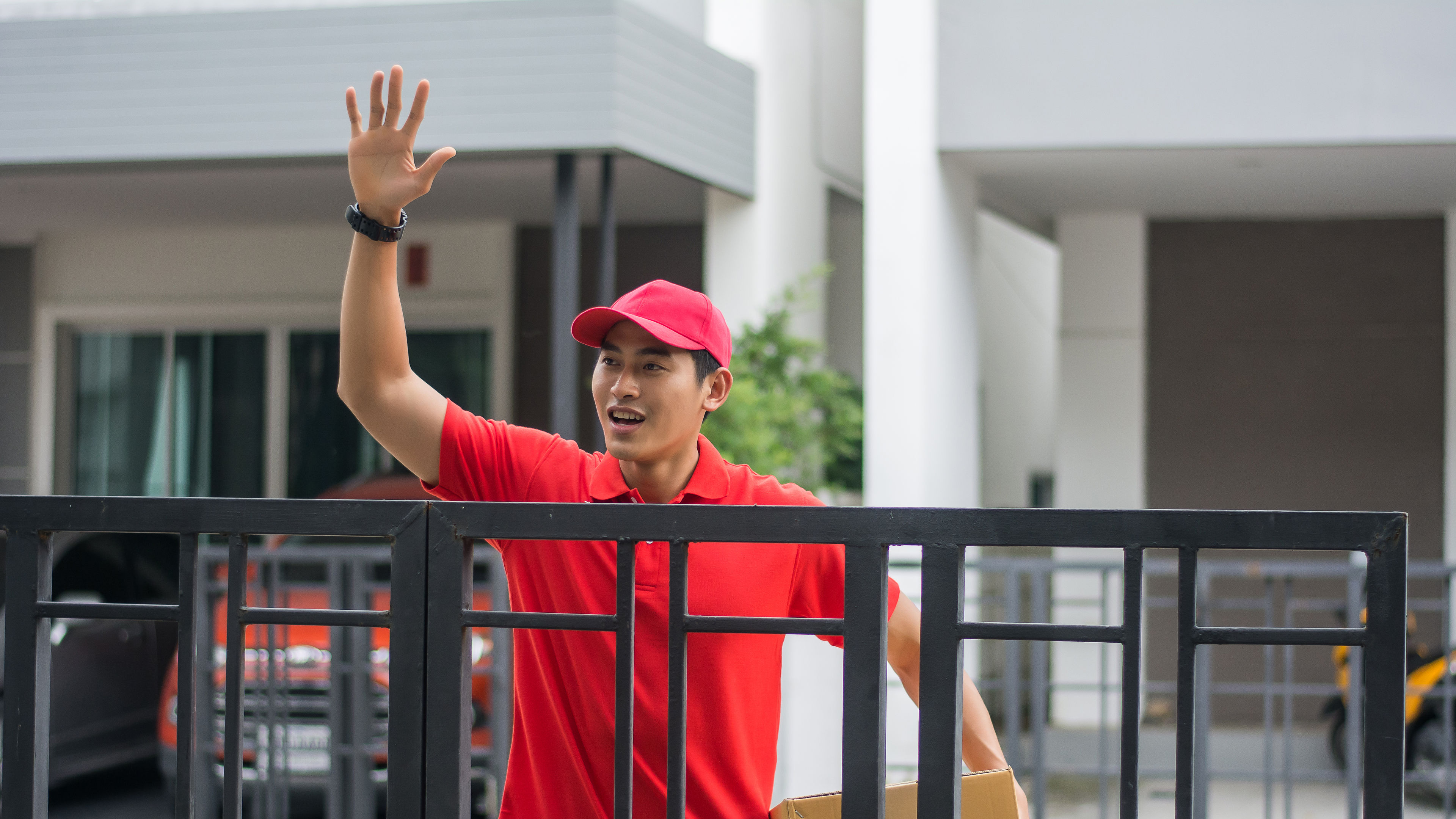 A delivery man with a carton in hand waving in the front of a home 
