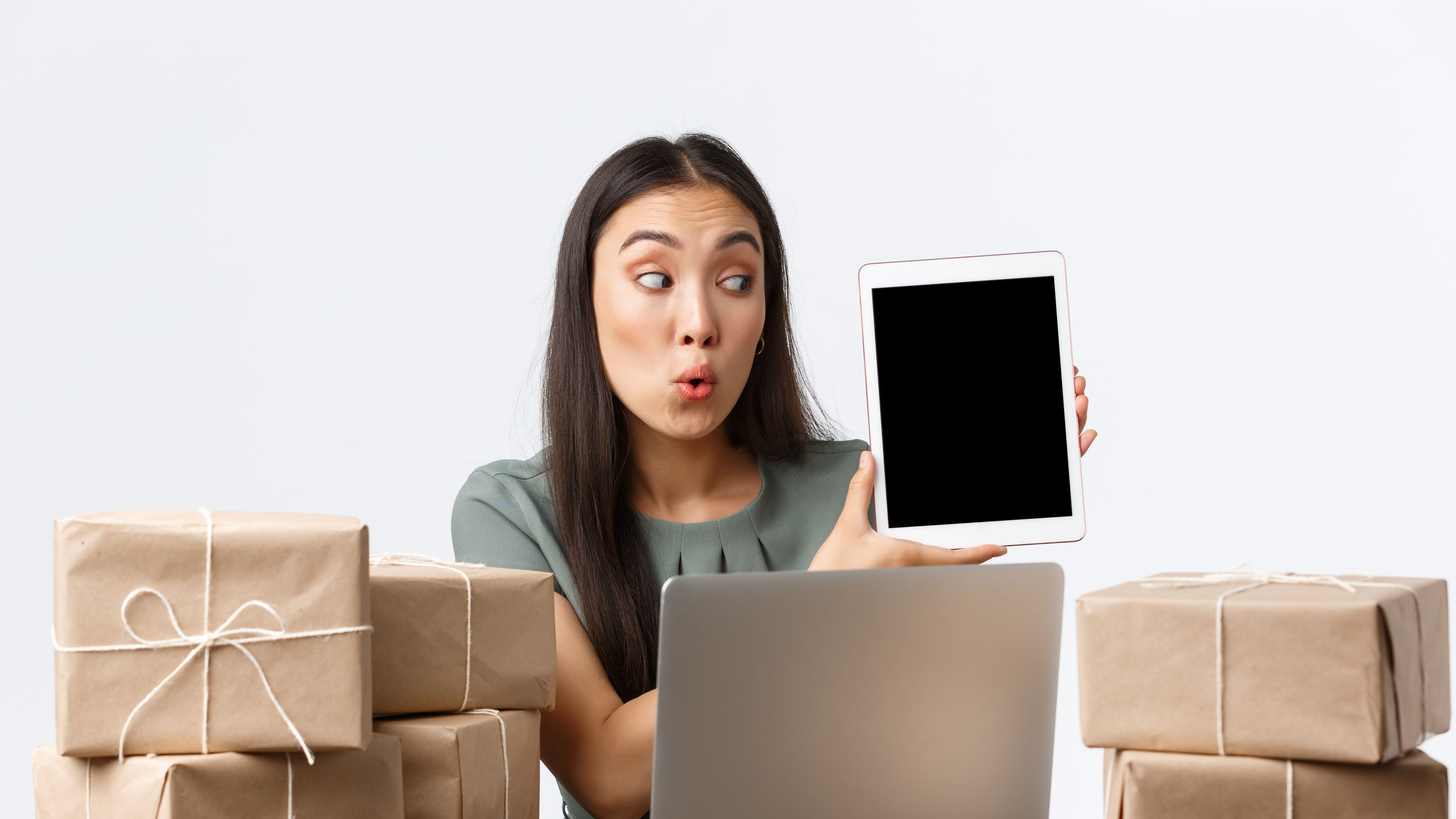 A woman holding a tablet and excitedly showcasing it while surrounded by wrapped packages and a laptop. 