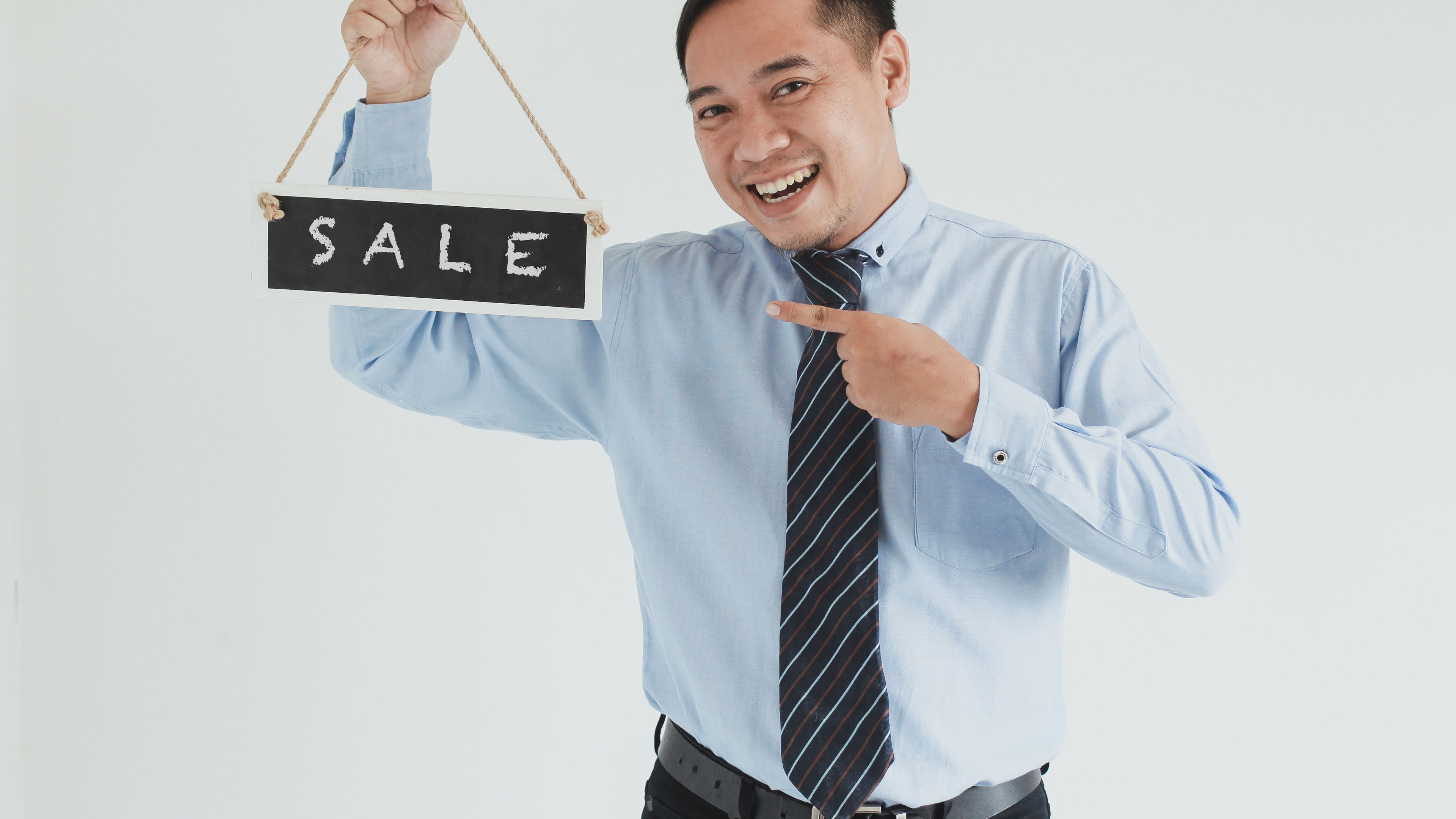 Happy sales man wearing blue shirt and tie posing with pointing and carrying "SALE" sign board on white background 