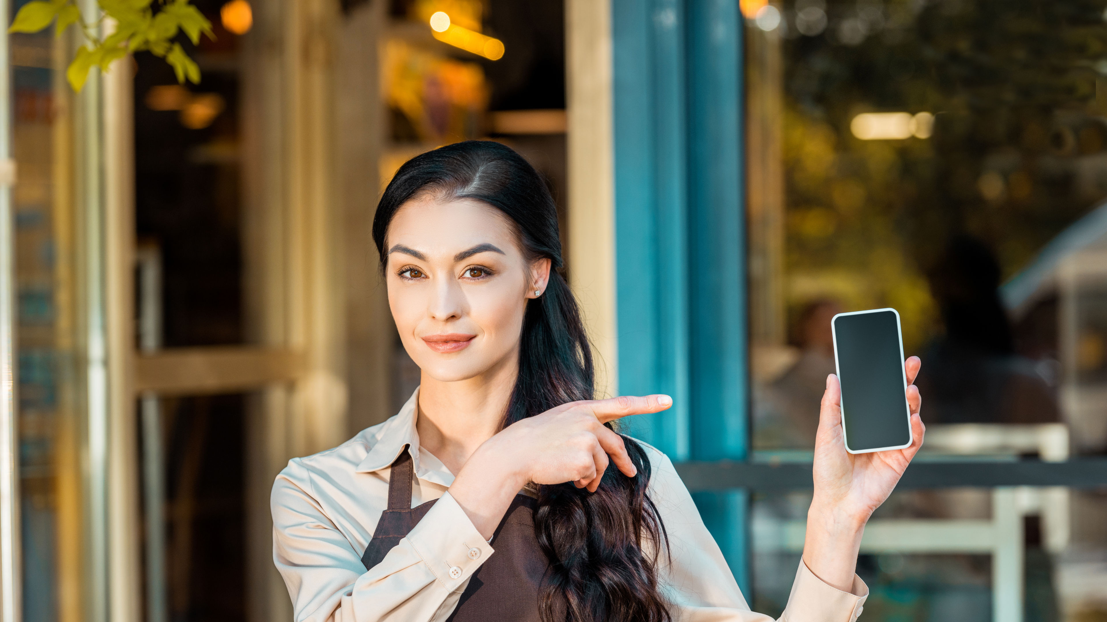 beautiful waitress in apron pointing on smartphone with blank screen  