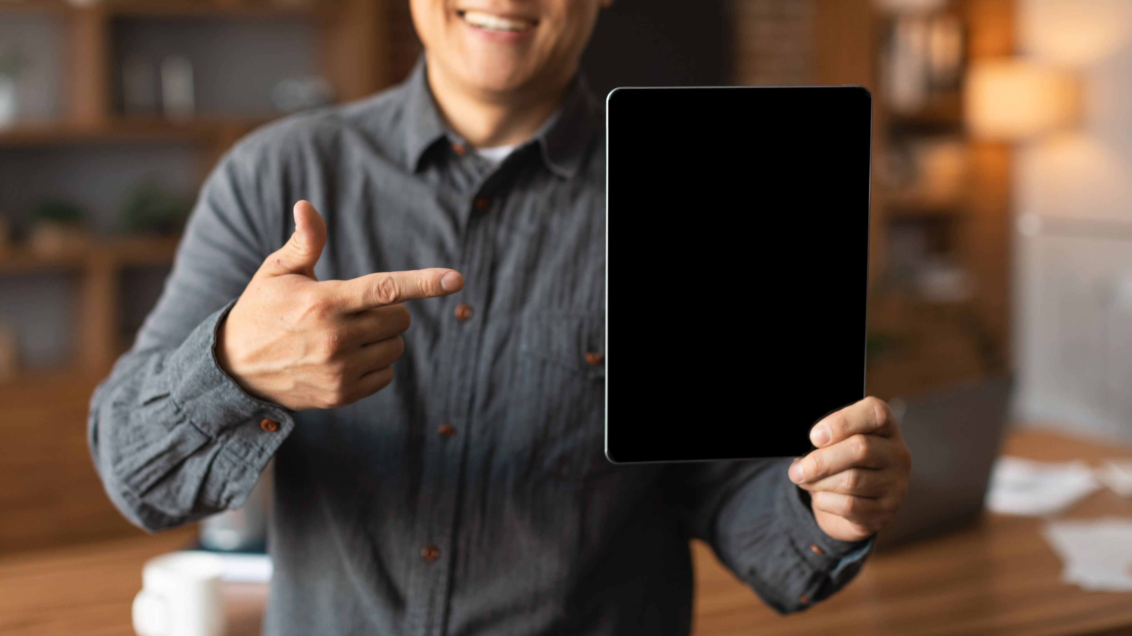 Cheerful adult chinese guy show finger at tablet with empty screen in office interior 
