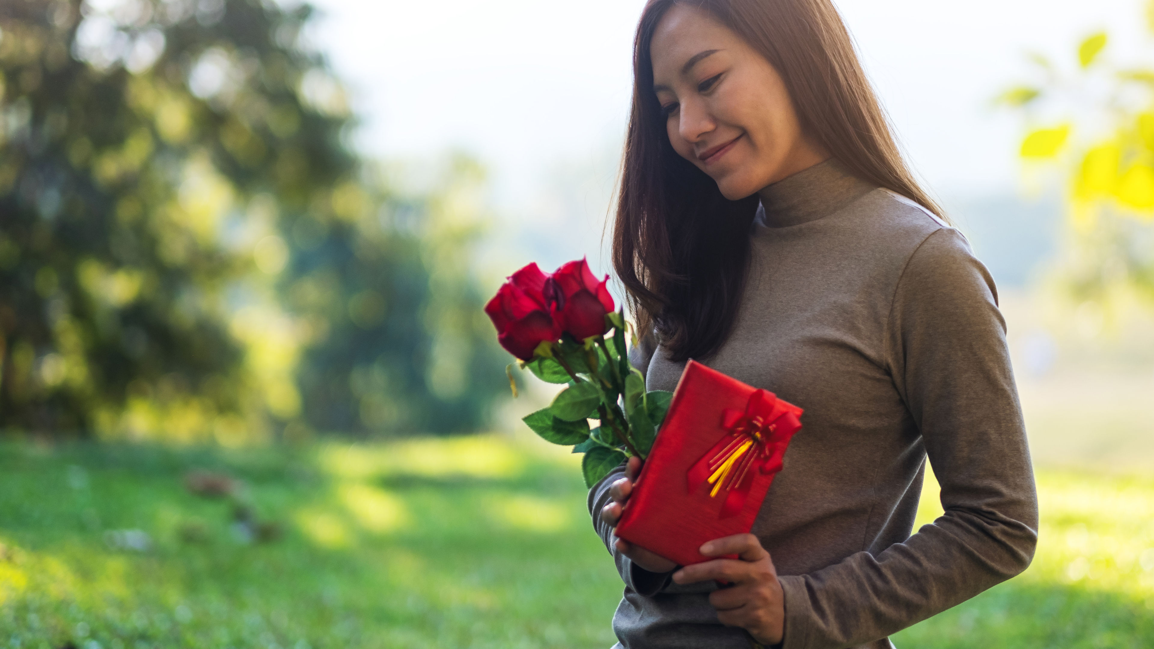 Closeup image of a beautiful young asian woman holding red roses flower and a gift box in the park 
