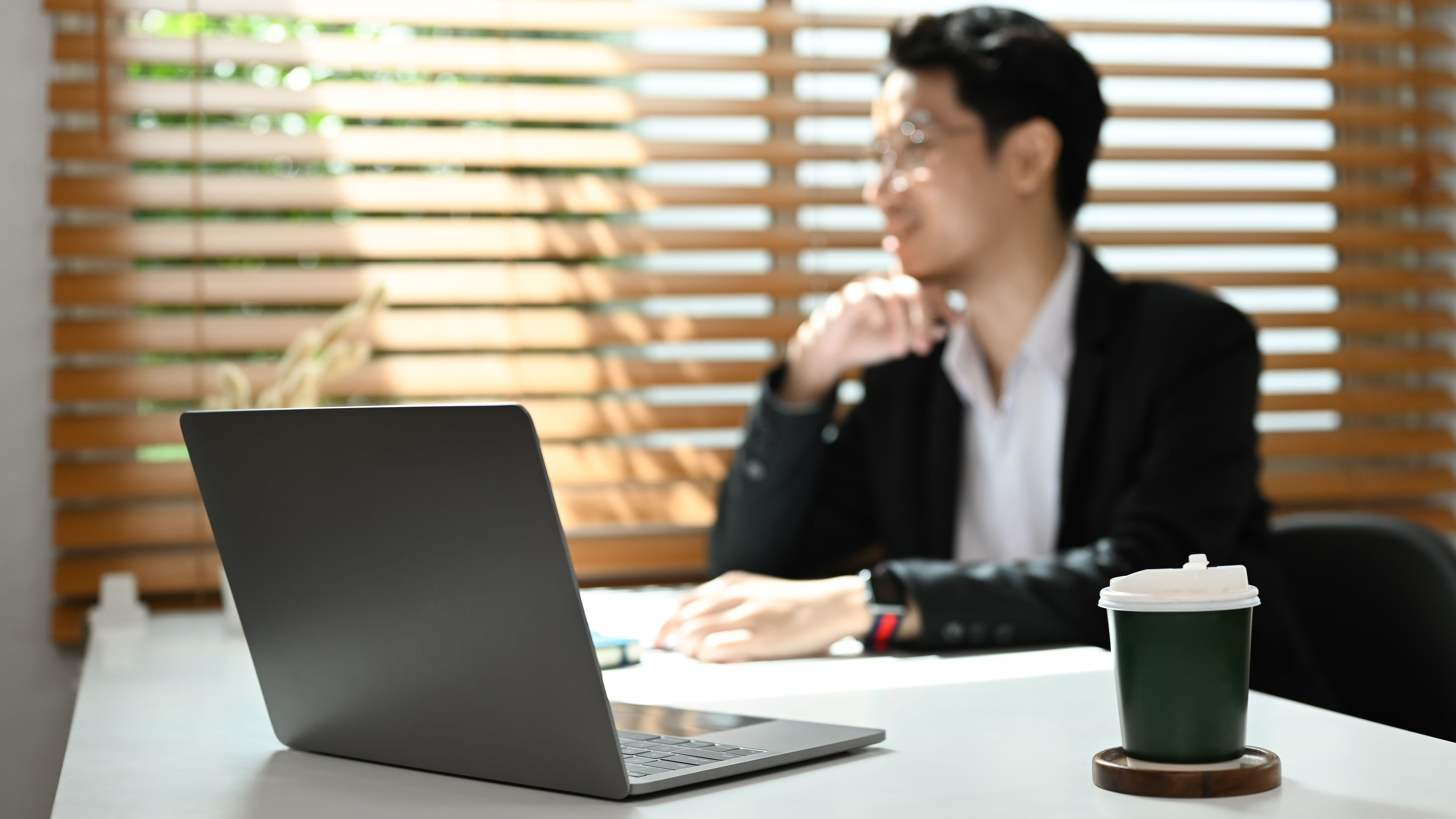 Laptop computer, paper cup of coffee on white table with thoughtful business man 
