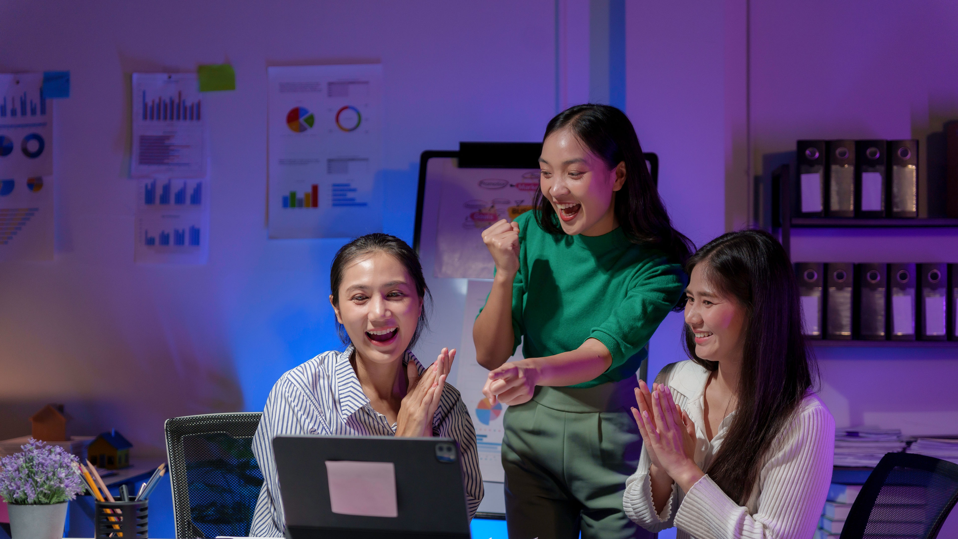 Three young businesswomen are looking excitedly at a laptop during a late night work session 