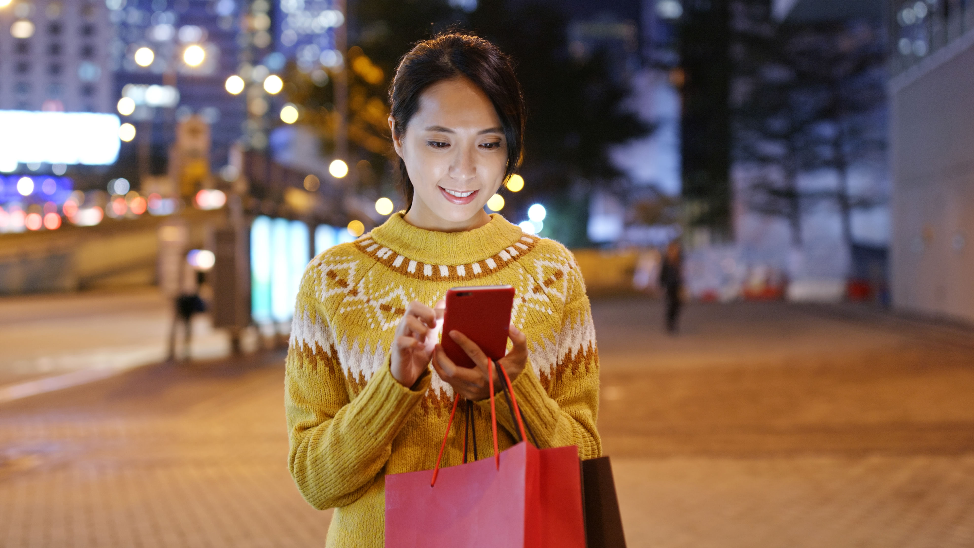 A woman wearing a cozy sweater holds shopping bags and browses her smartphone under city lights at night 