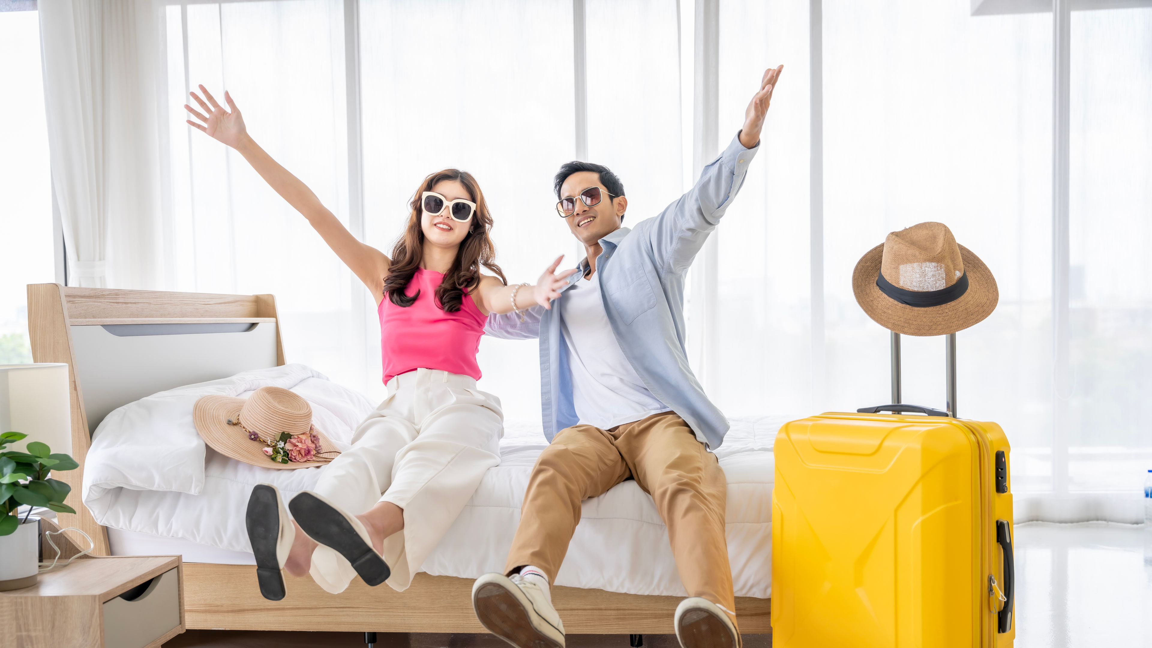 A cheerful couple sitting on a bed with open arms, a yellow suitcase and hats nearby, hinting at travel.