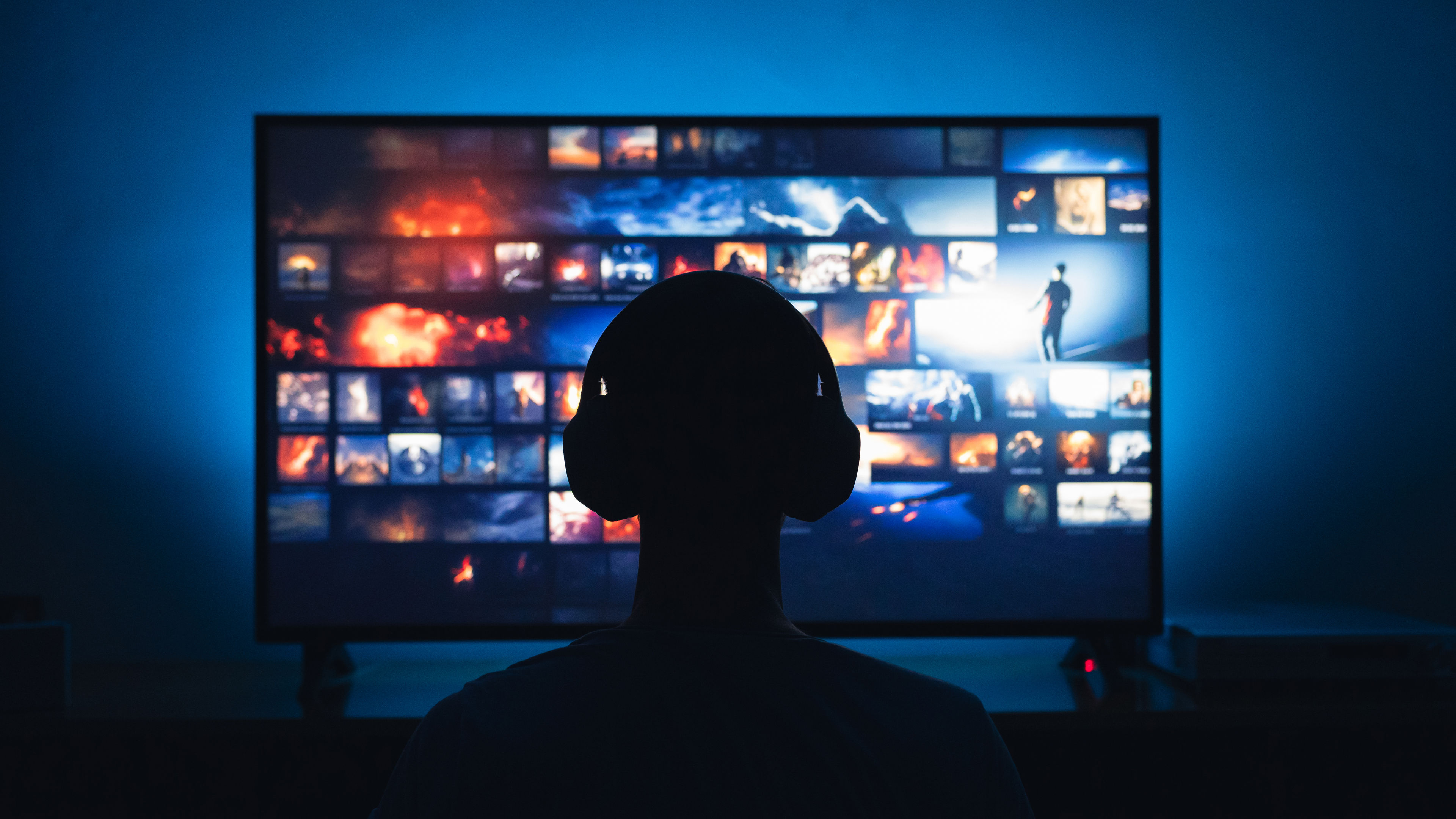 A man with headphones on, sitting in front of a TV screen, watching intently.