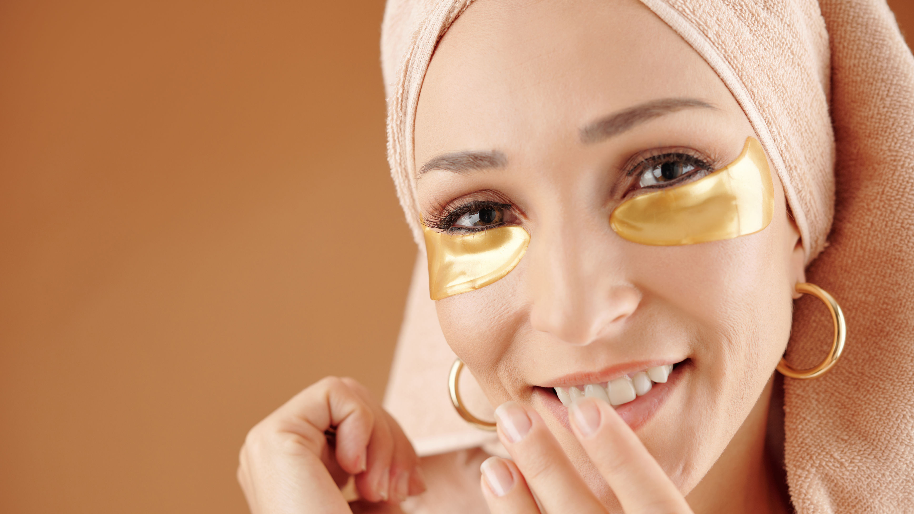 A woman with an under-eye mask on, smiling warmly as she relaxes and enjoys her self-care routine.