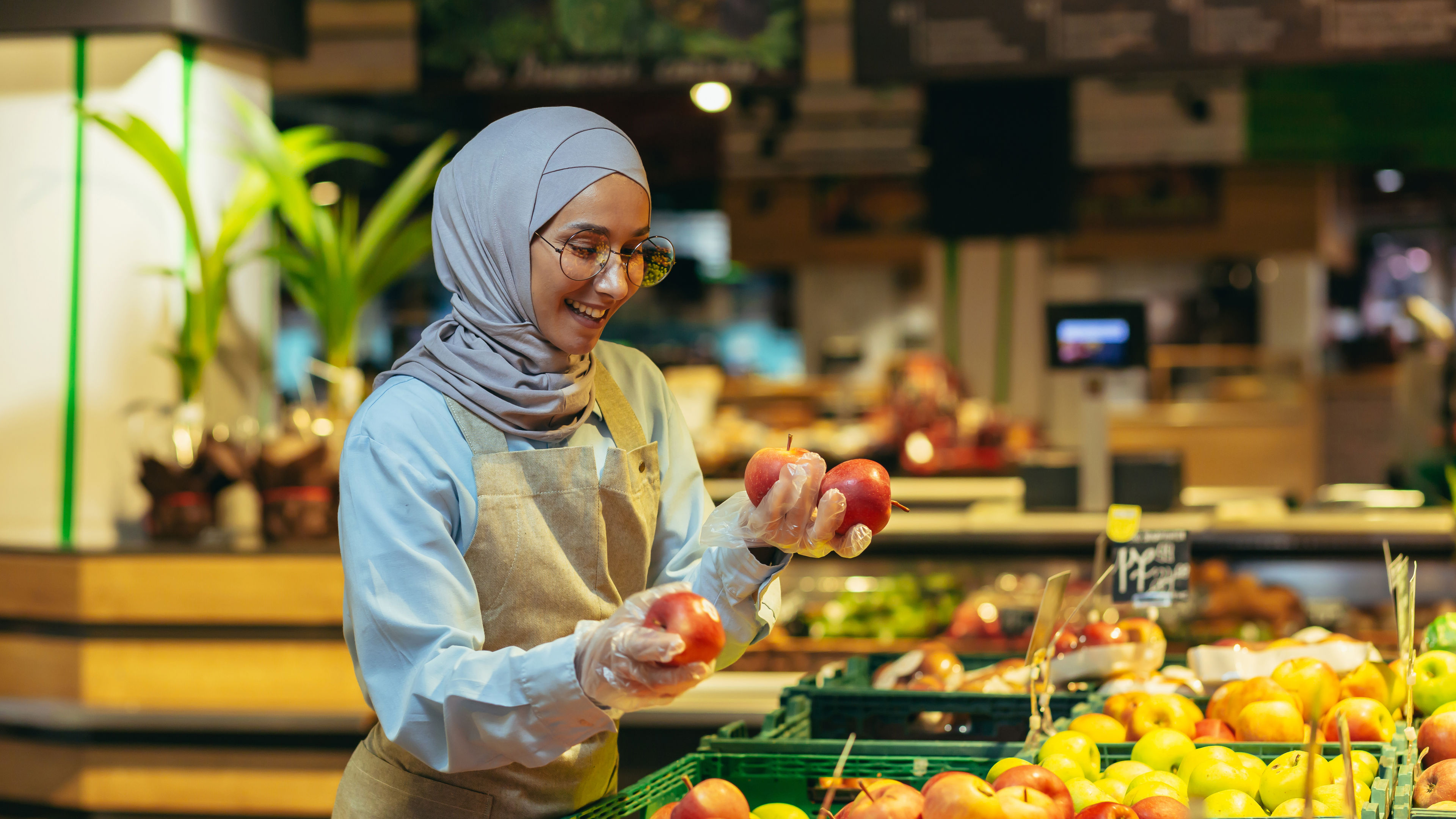 بائعة ترتدي الحجاب ترتدي مئزرا وتبتسم أثناء تصفح التفاح وتطلقه في السوبر ماركت. 