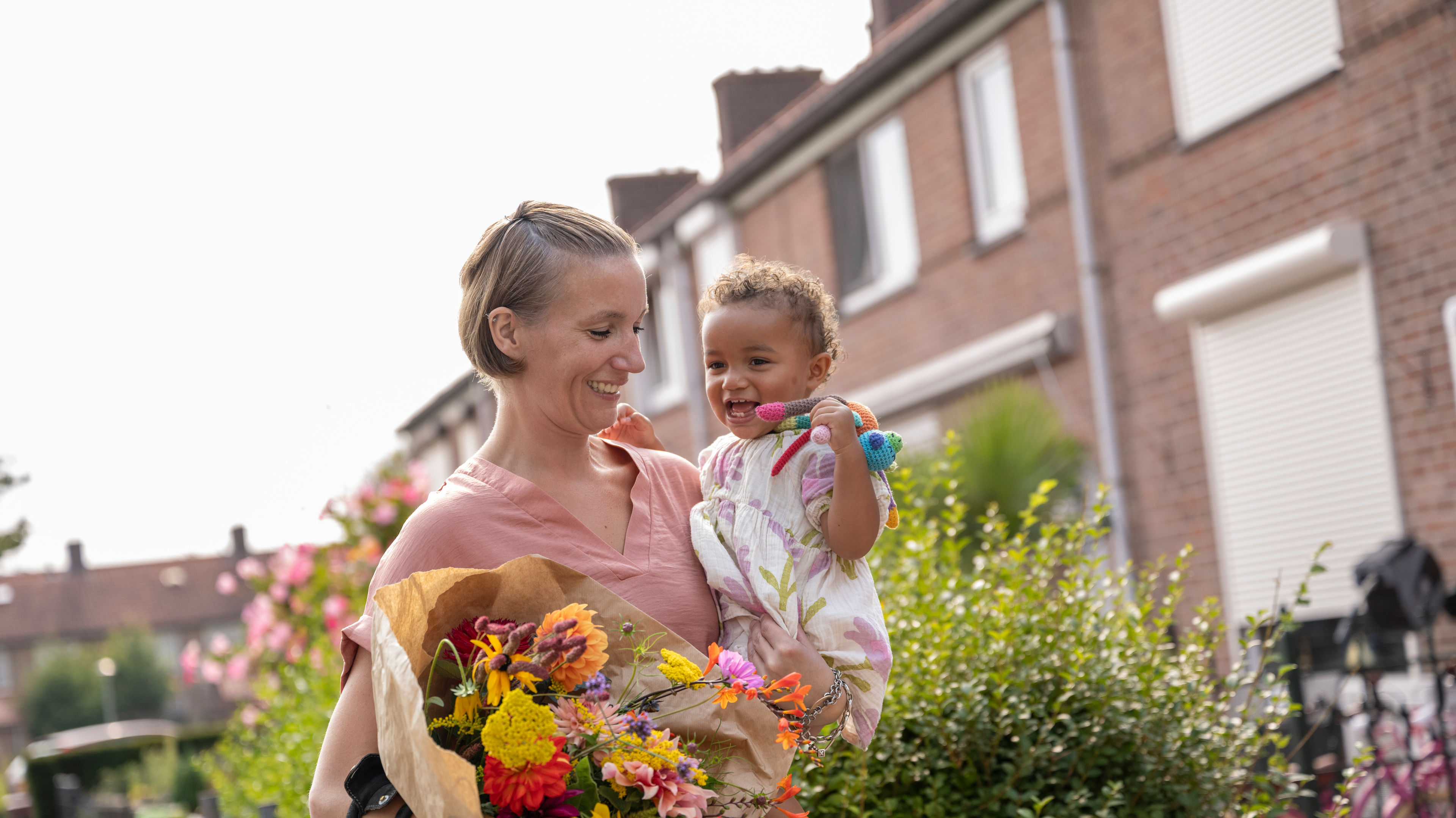 A smiling woman carrying her cheerful toddler and a bouquet of flowers while walking outdoors 