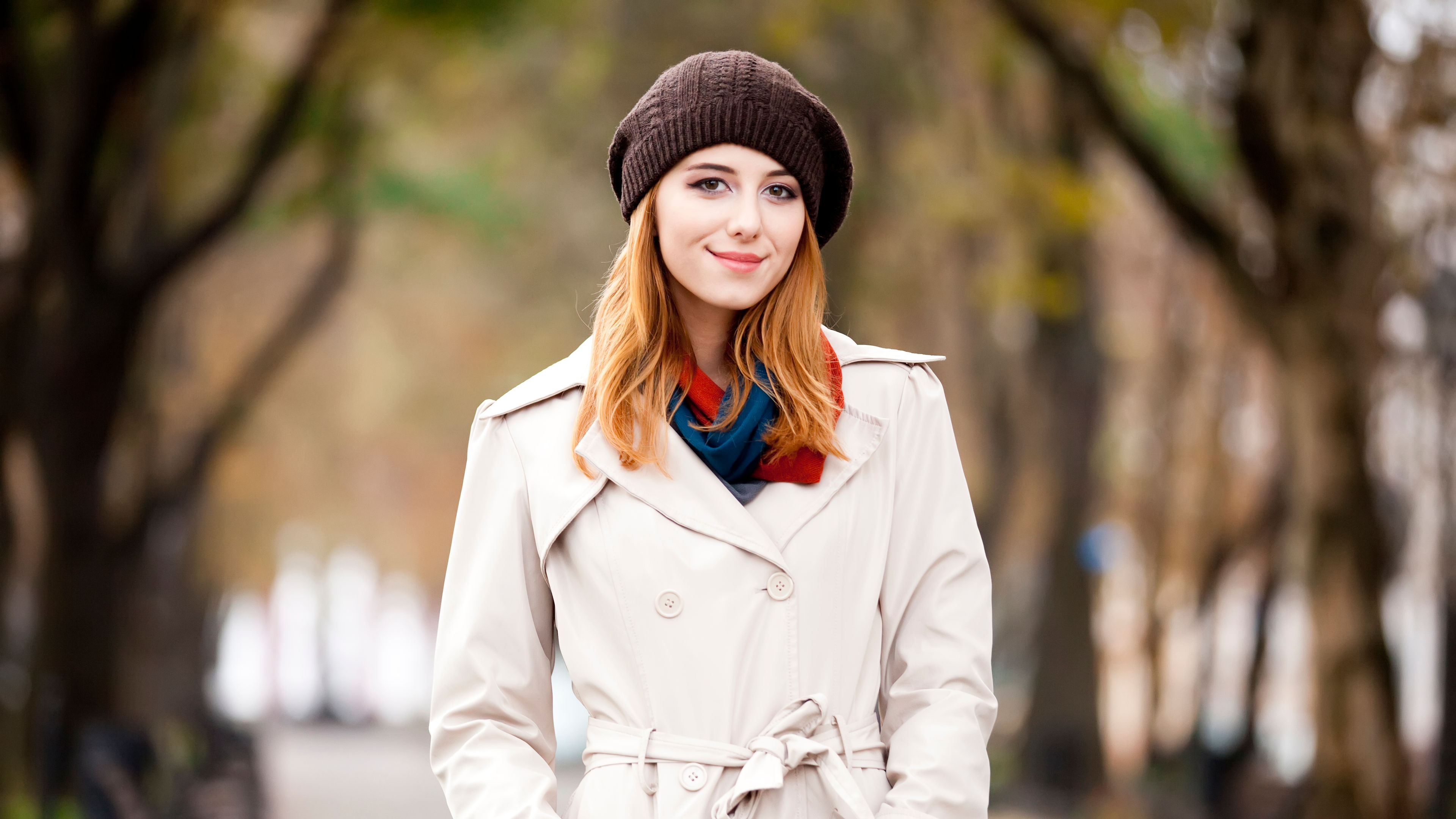 a young woman wearing a beige trench coat, a brown knit hat, and a colorful scarf 