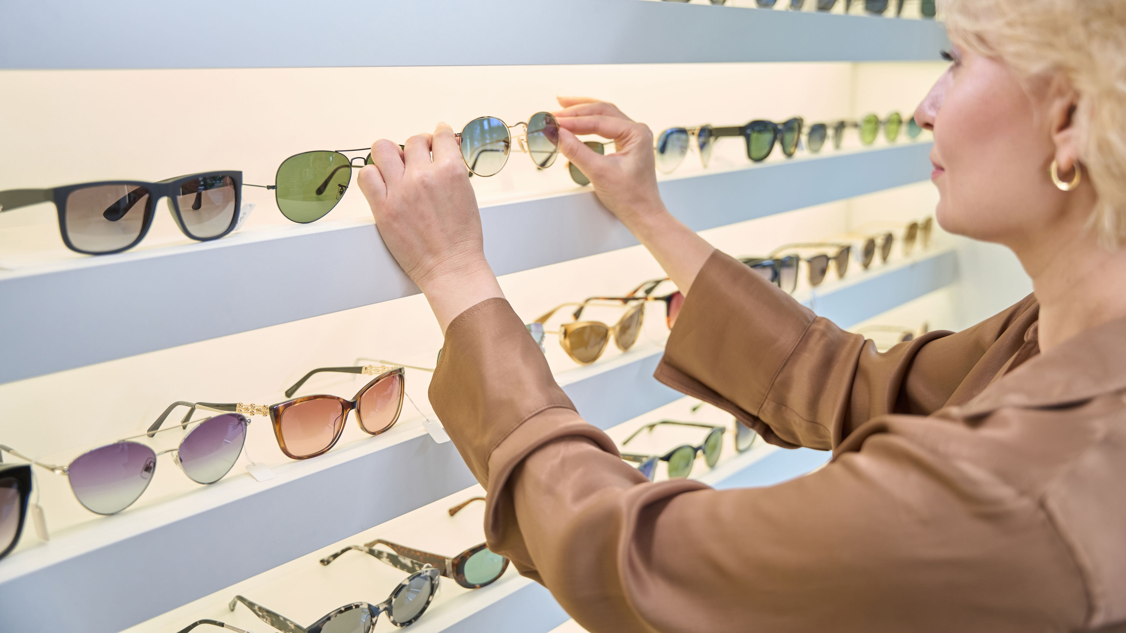 A woman wearing a light brown blouse, carefully choosing a pair of sunglasses