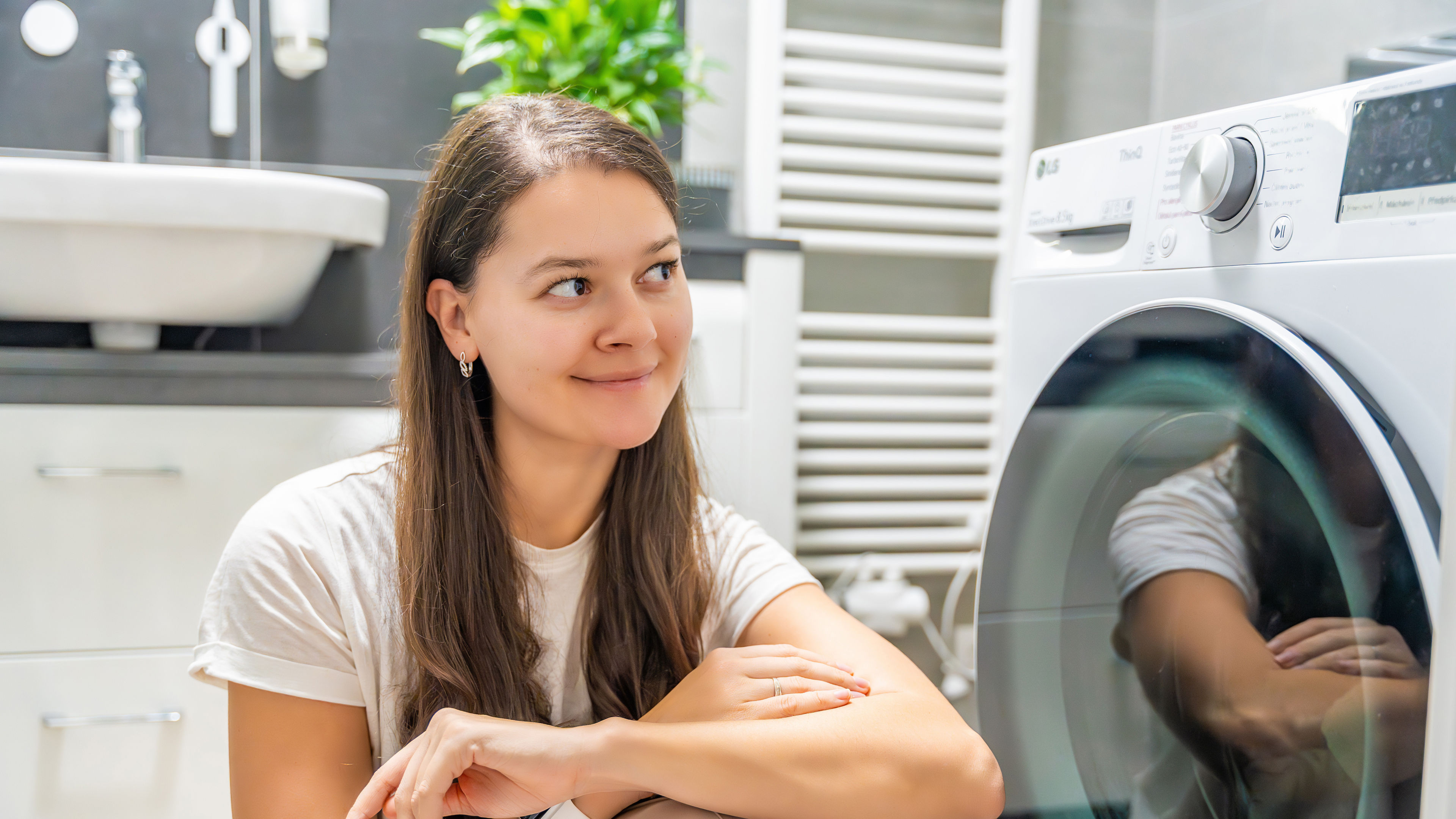 A young woman sits nearby, staring intently at the washing machine door as she patiently waits for the laundry cycle to finish in a clean 