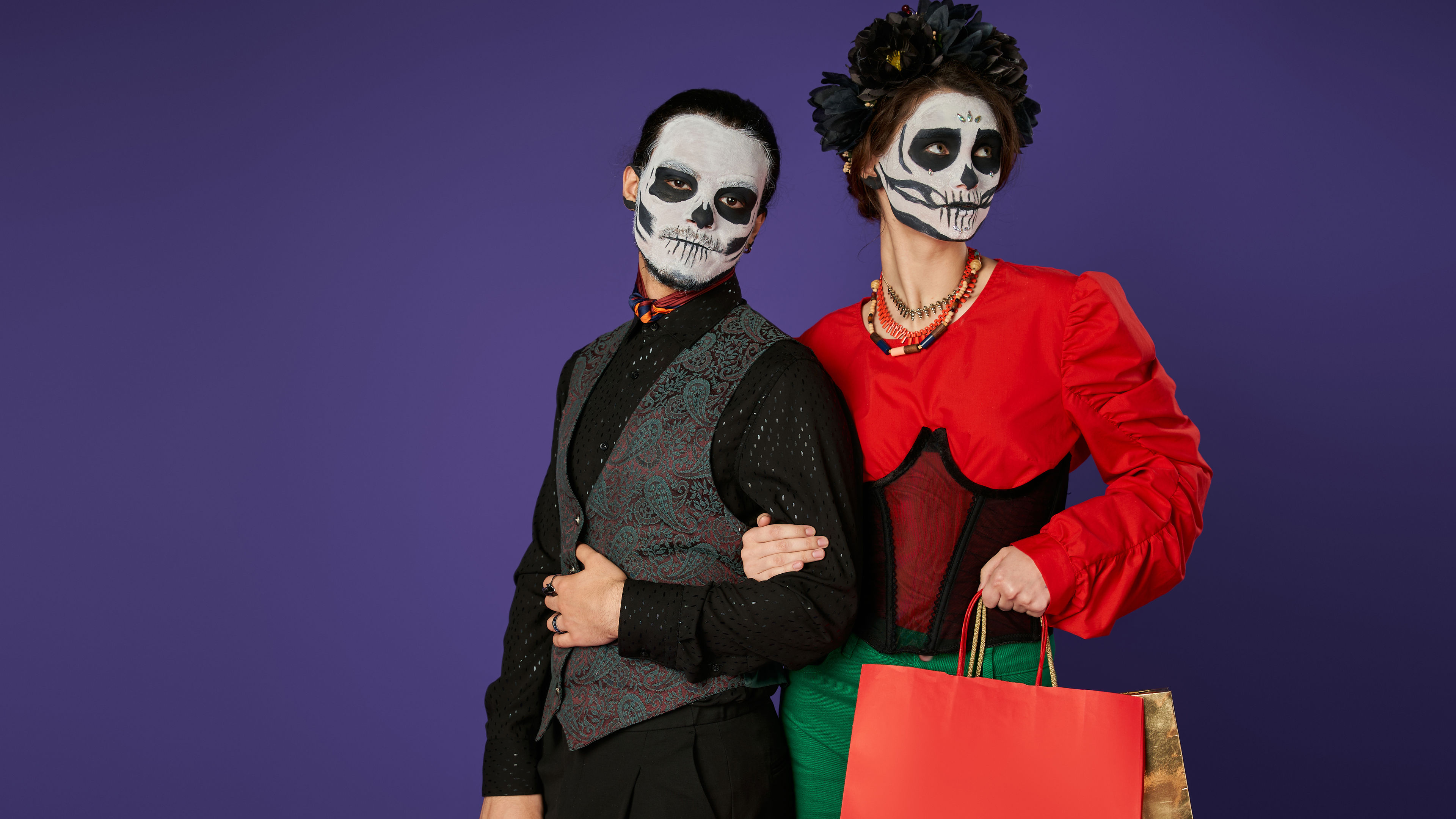 An elegant couple with skull face paint dressed in festive costumes, posing together while holding shopping bags 