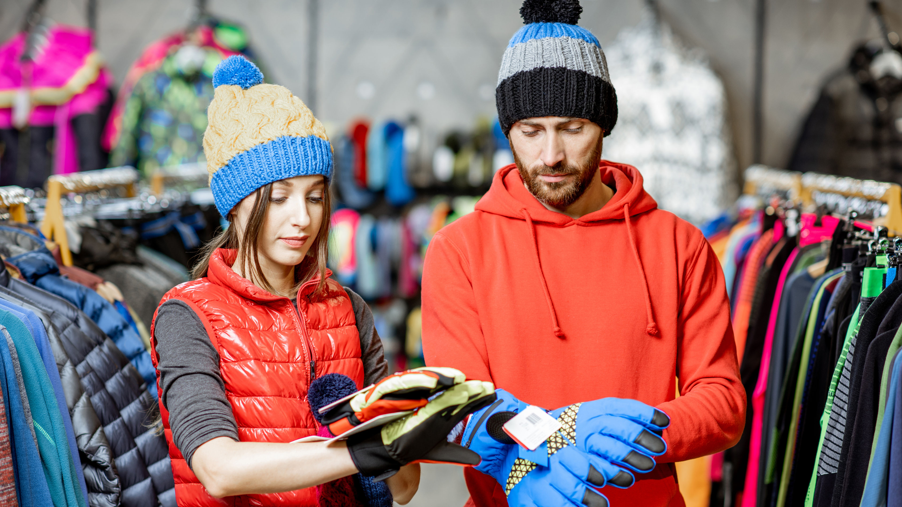 Young couple trying winter clothes wearing colorful hats and gloves in the sports shop 
