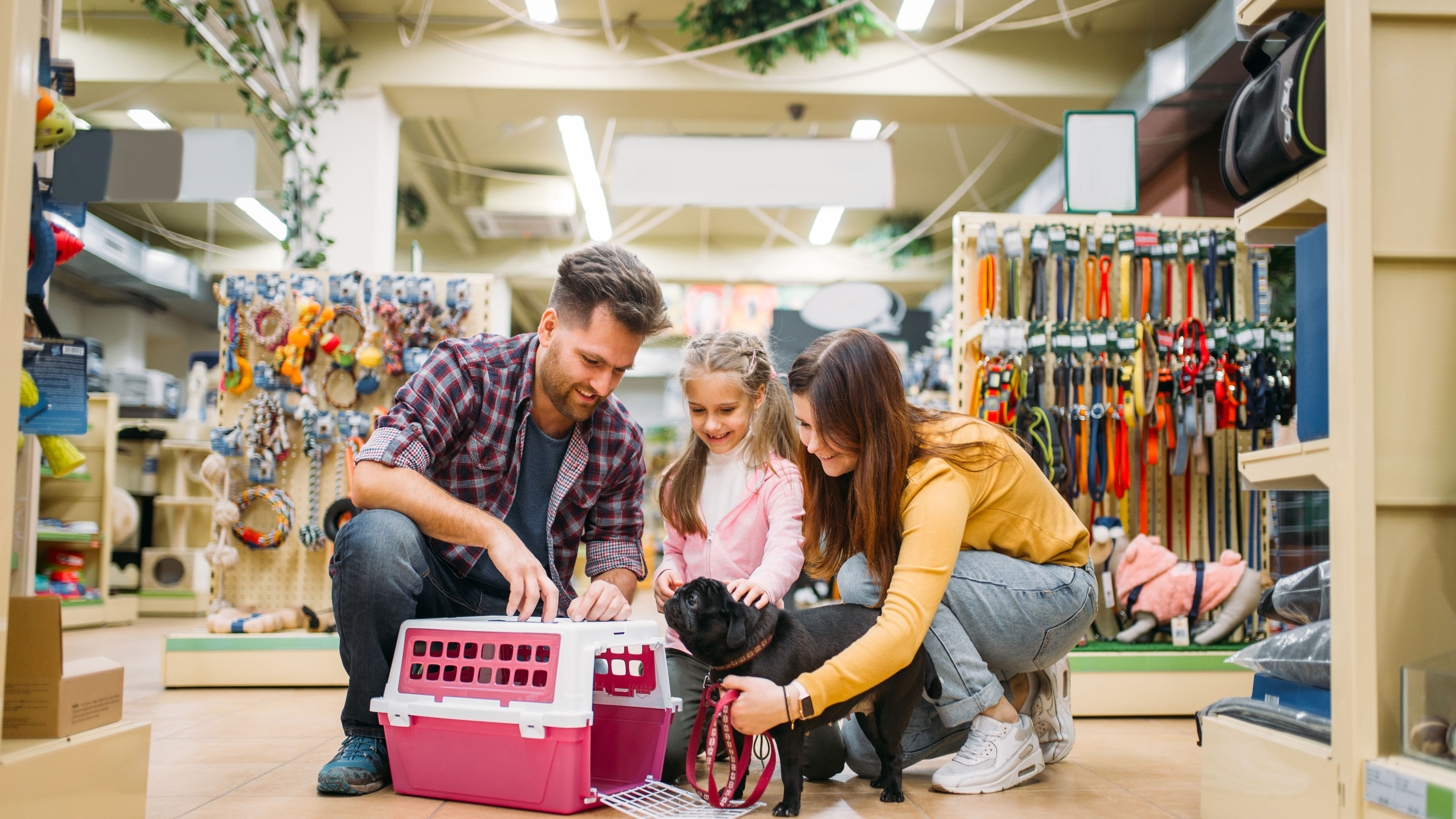 A cheerful family of three kneels in a pet store, browsing supplies for their new small black dog 