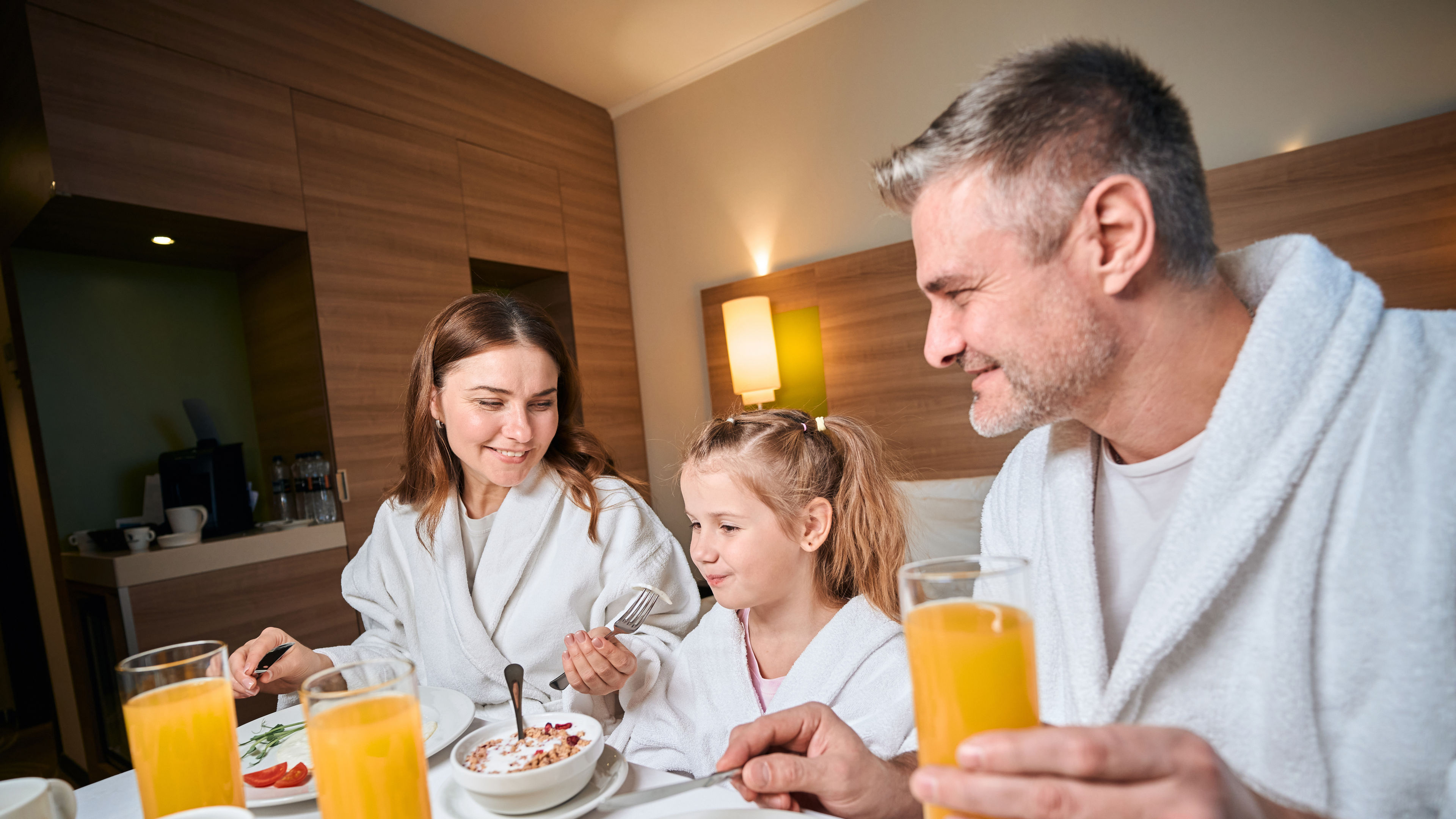 Parents and a child wearing robes enjoy a cheerful breakfast in a hotel room, with food and orange juice on the table. 