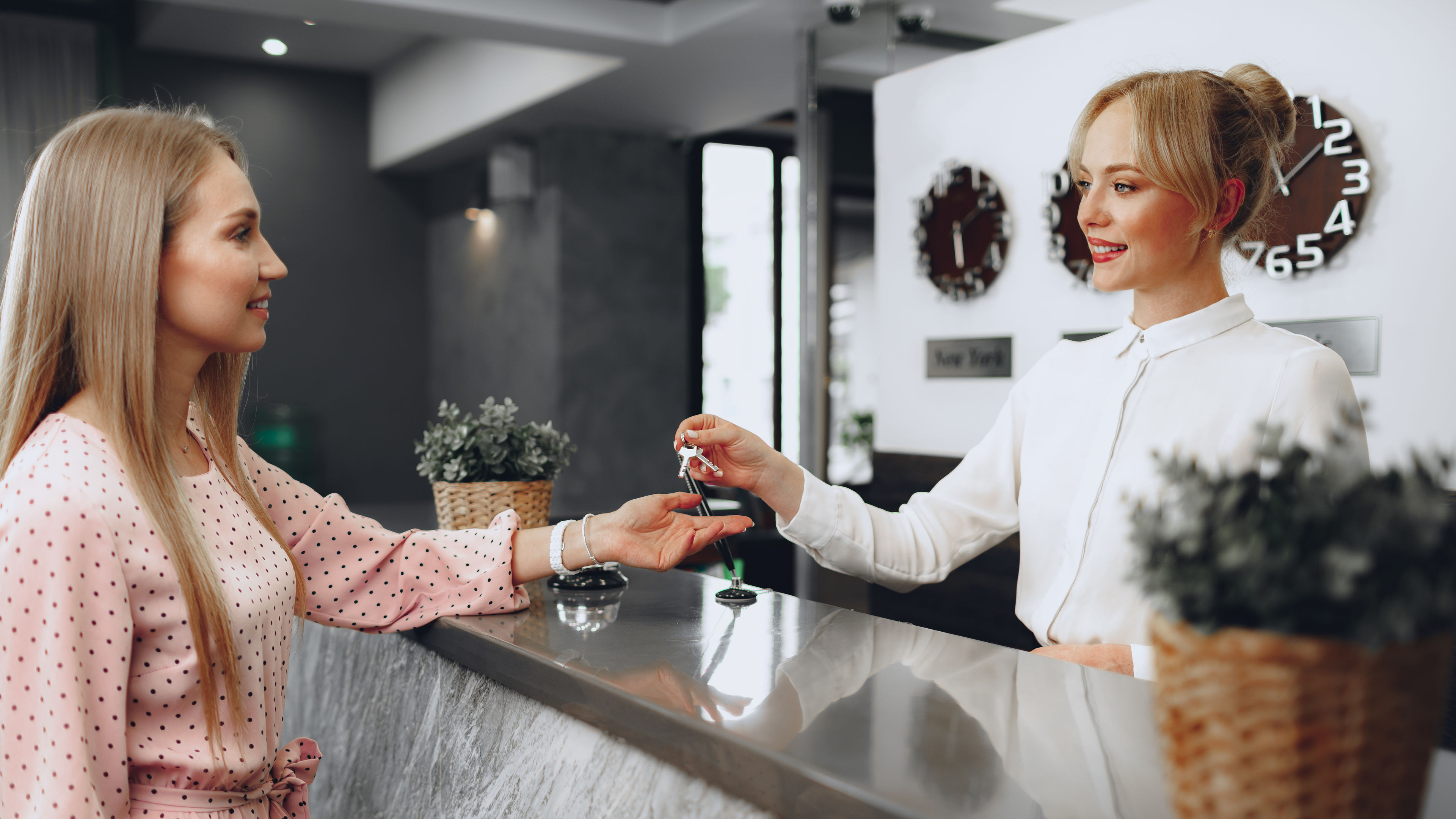 A hotel receptionist hands a key card to a woman in a pink polka-dot dress at a modern check-in desk 