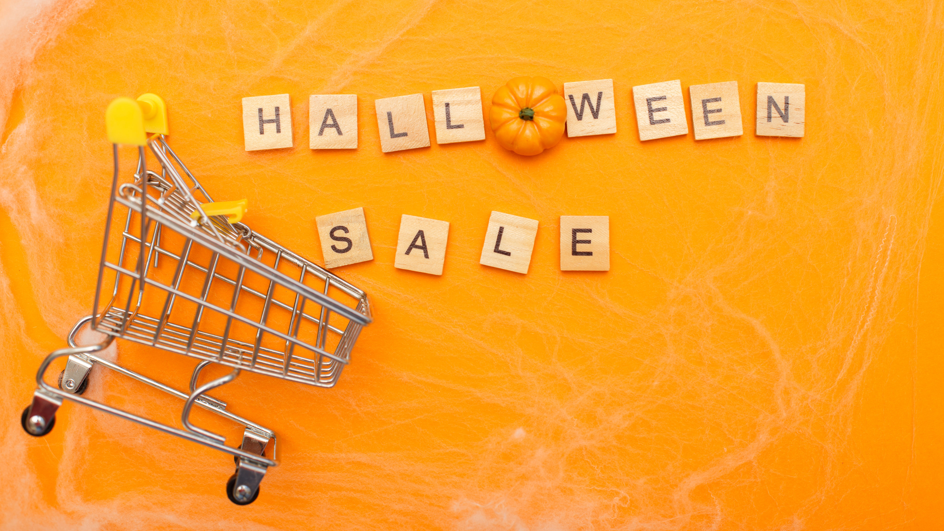 A small shopping cart on a bright orange background with cobwebs, featuring scrabble tiles spelling "HALLOWEEN SALE" and a miniature pumpkin. 