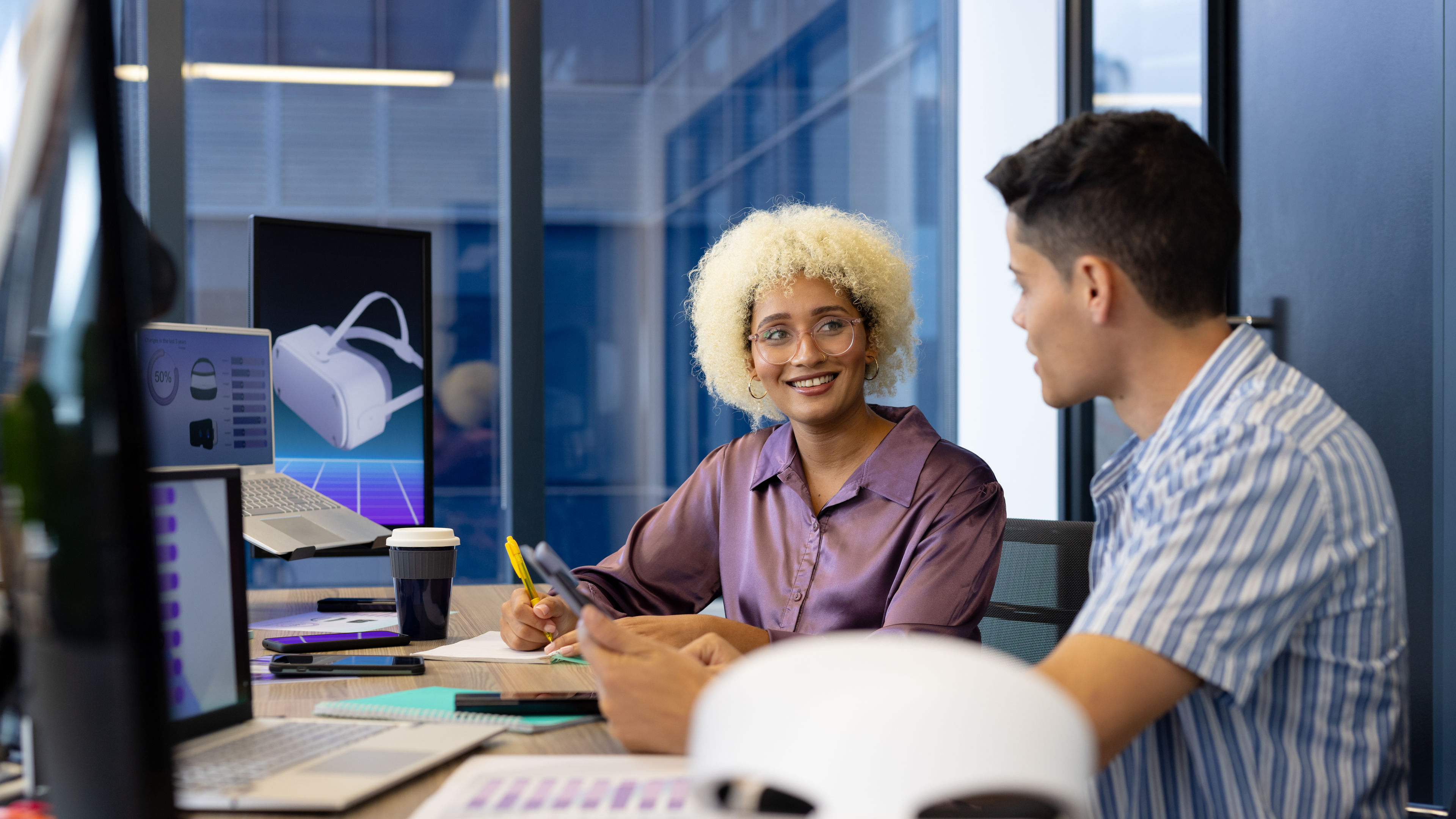 In modern office, diverse young team wearing glasses, discussing. She, with curly blonde hair, examining something on tablet.