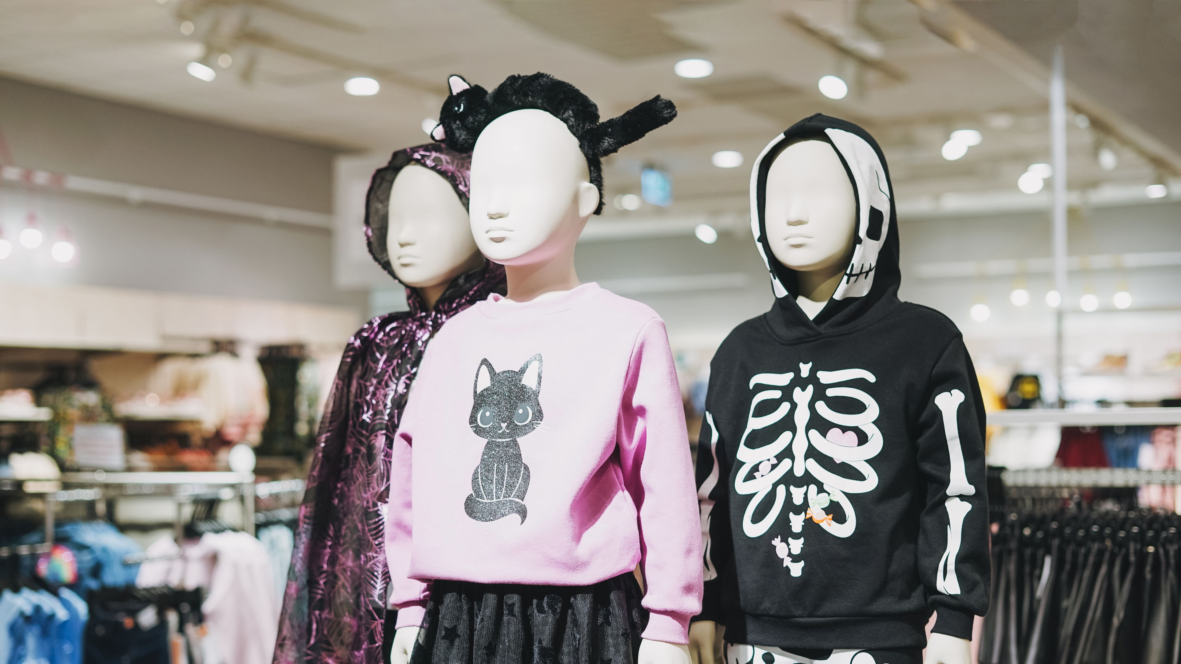 A display of children’s Halloween costumes on mannequins, featuring skeleton, cat, and witch-themed outfits in a store setting 